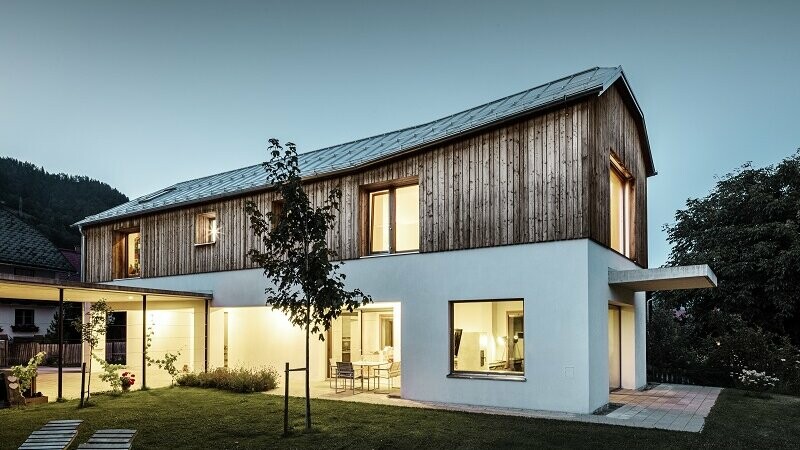 Frontalansicht des Einfamlienhauses im Ort Pruggern bei Dämmerung. Im Haus brennt Licht. Das Dach wurde mit PREFALZ in der Farbe Naturblank eingedeckt.