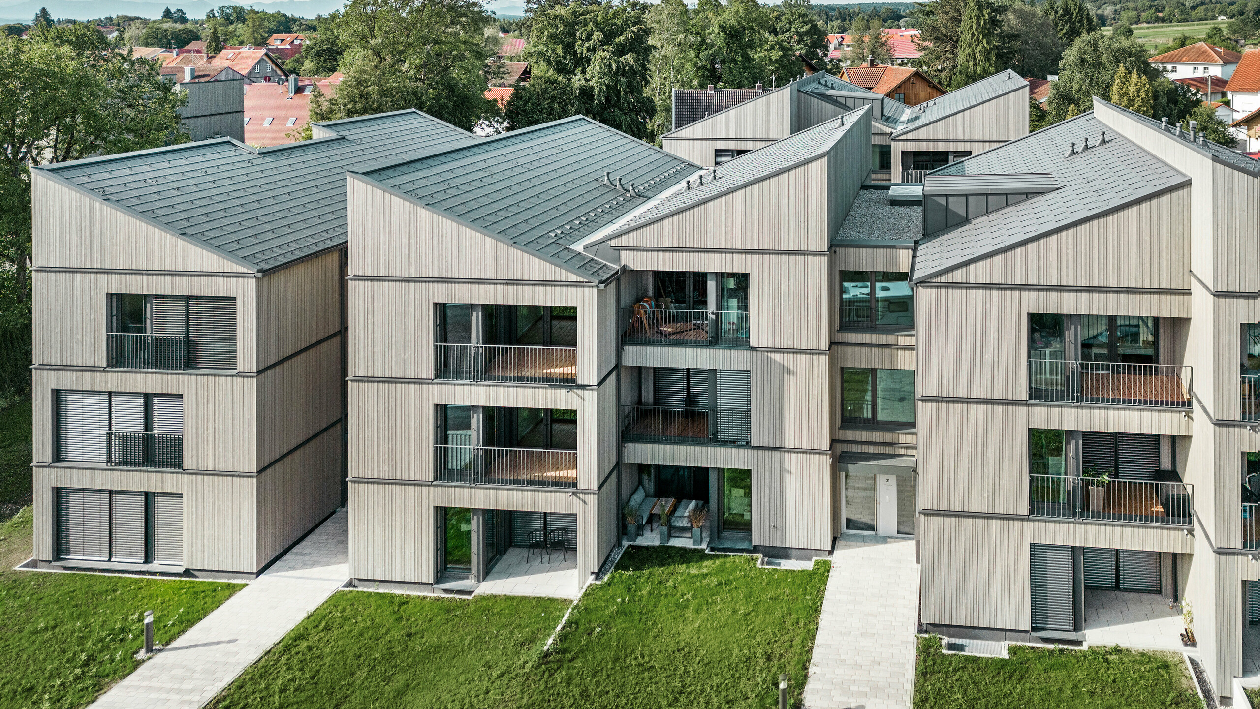 Luftaufnahme des modernen Mehrfamilienhauses am Schmucker-Areal in Utting am Ammersee, mit nachhaltiger Holzhybridbauweise und eleganten Holzfassaden. Die Gebäude sind mit langlebigen PREFA Dachplatten R.16 in P.10 Dunkelgrau ausgestattet. Diese innovative Architektur, geplant von WWA Architekten aus München, zeichnet sich durch klare Linien und harmonische Integration in die grüne Umgebung aus. Die großzügigen Balkone und großen Fenster sorgen für eine helle und freundliche Wohnatmosphäre, während die hochwertigen Materialien für Langlebigkeit und Wetterbeständigkeit sorgen.