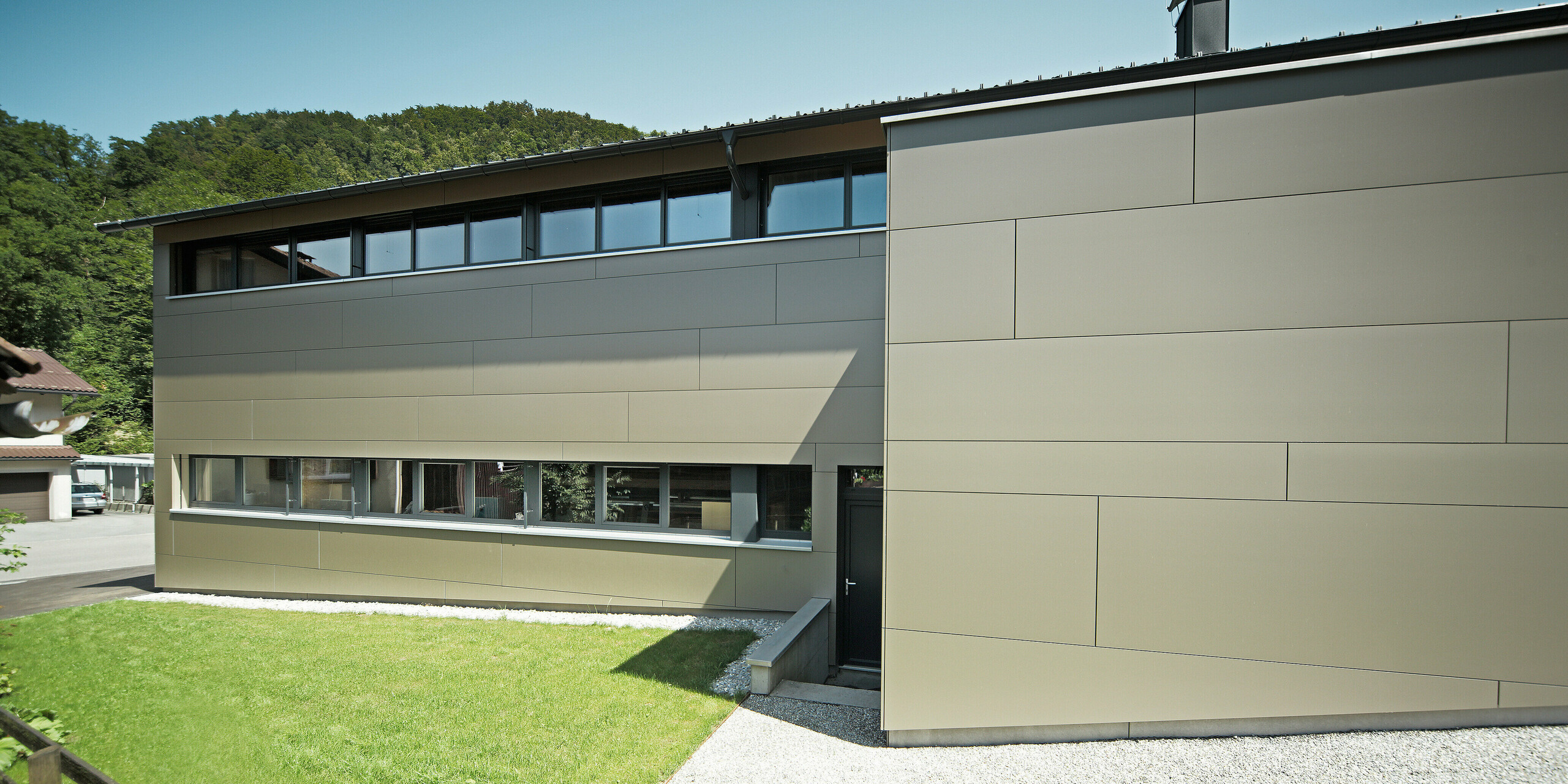 Ansicht eines Einfamilienhauses von der Gartenseite in Hohenems, verkleidet mit PREFA Aluminium Verbundplatten in der Farbe Bronzemetallic. Die harmonische Verbindung von moderner Architektur und Natur wird durch die große Fensterfront und die klaren Linien der Fassadenplatten unterstrichen, während die PREFA Dachplatten in P.10 Anthrazit für eine optische Einheit sorgen und das Haus vor den Elementen schützen.