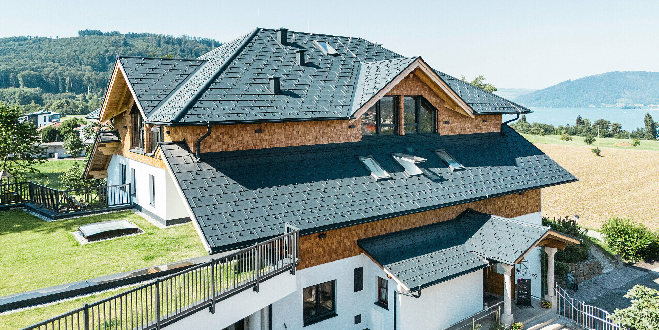 Vogelperspektive auf das Café-Restaurant Zenz'n Stub'n in Schörfling am Attersee, das mit PREFA Dachplatten R.16 in P.10 Anthrazit neu eingedeckt wurde.Das langlebige Aluminiumdach verleiht dem Gebäude ein modernes und stilvolles Erscheinungsbild und bietet gleichzeitig robusten Schutz. Die harmonisch integrierte Dachentwässerung und die natürliche Umgebung runden das Gesamtbild ab.