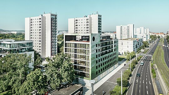 Luftaufnahme des Wohn- und Geschäftshauses "Pick-Nick" am Straßburger Platz in Dresden mit der markanten PREFA Fassade aus Aluminiumrauten in verschiedenen Grün- und Beigetönen. Eingebettet in ein modernes urbanes Umfeld mit Hochhäusern und Grünflächen liegt das Gebäude an einer Hauptverkehrsstraße. Die prägnante Architektur und die hochwertige PREFA Fassade setzen optische Akzente im Stadtbild.