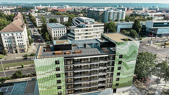 Luftaufnahme des Wohn- und Geschäftshauses "Pick-Nick" in Dresden mit einer PREFA Fassade aus Wandrauten in verschiedenen Grün- und Beigetönen. Im Hintergrund ist das beeindruckende Stadtpanorama zu sehen. Das Gebäude verfügt über eine großzügige Dachterrasse mit Blumenbeeten und einem Kinderspielplatz. Eingebettet in ein urbanes Umfeld bietet es eine Mischung aus moderner Architektur, Nachhaltigkeit und funktionalem Design.