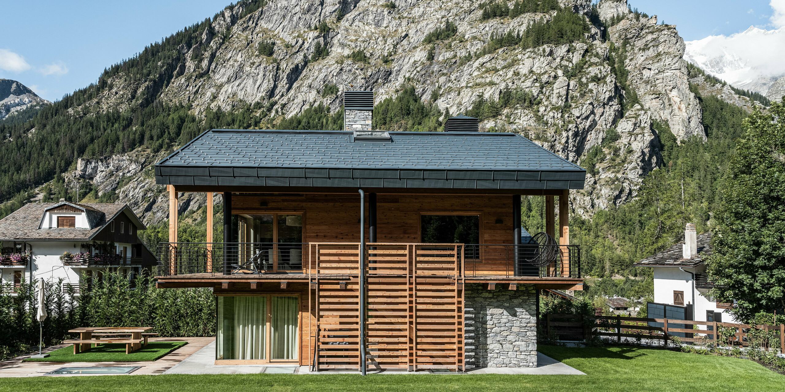 Idyllische Villa in Courmayeur mit einer harmonischen Kombination aus Holz, Stein und Aluminium an der Fassade und am Dach. Die PREFA FX.12 Dach- und Fassadenpaneele in P.10 Steingrau präsentieren sich vor einer beeindruckenden Bergkulisse, die die alpine Atmosphäre unterstreicht. Markante Details wie das großzügige Balkongeländer und die großen Fenster sorgen für eine offene und einladende Atmosphäre, während der gepflegte Garten zum Entspannen im Freien einlädt.