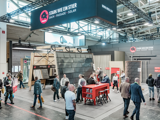 Der PREFA Messestand auf der BAU Messe, gekennzeichnet durch den Slogan „Stark wie ein Stier – Dach, Fassade, Solar“ und eine große hängende Werbefläche. Im Zentrum stehen präsentierte Aluminium-Dach- und Fassadensysteme sowie innovative Montagelösungen. Besucher tauschen sich mit PREFA Mitarbeitern an roten Beratungstischen aus, während weitere Exponate und Infotafeln im Hintergrund zu sehen sind.