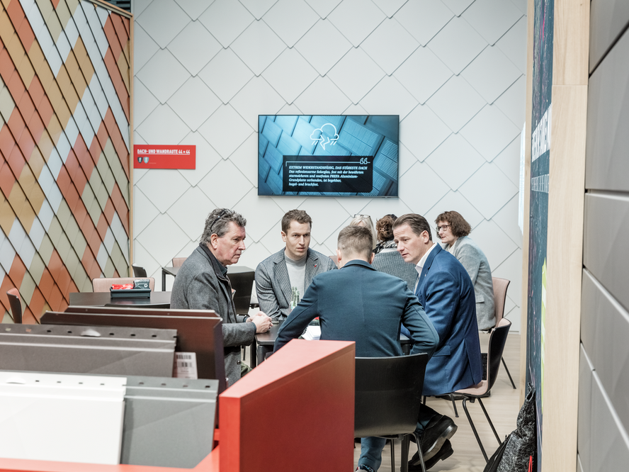 Fachgespräch am PREFA Messestand auf der BAU Messe. Mehrere Personen sitzen an einem Tisch im Beratungsbereich, umgeben von Musterflächen mit Wandrauten in verschiedenen Farbtönen. Ein Bildschirm im Hintergrund zeigt den PREFA Messefilm.