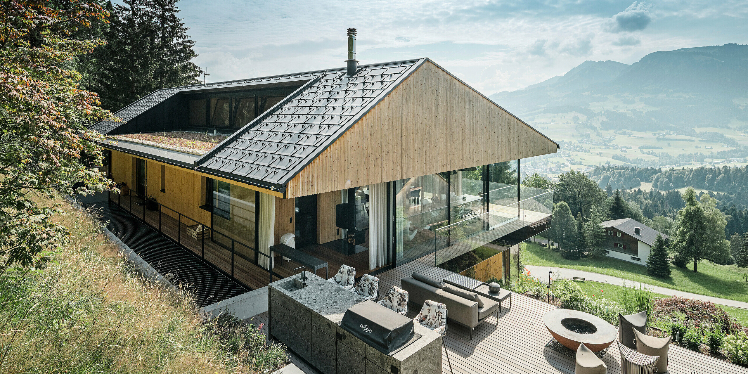 Einfamilienhaus in Egg, Vorarlberg, mit PREFA Dachplatten R.16 in P.10 Schwarz und beeindruckendem Bergpanorama im Hintergrund. Die hochwertige Dachkonstruktion aus Aluminium wird durch einen effizienten Schneeschutz ergänzt.Im Vordergrund die großzügige Terrasse mit moderner Aussenküche und stilvollen Gartenmöbeln. Die Möblierung fügt sich harmonisch in die natürliche Umgebung ein.