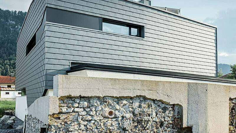 Fassadenverkleidung mit der PREFA Wandschindel aus Aluminium in Hellgrau mit einer Steinmauer vor dem Haus;