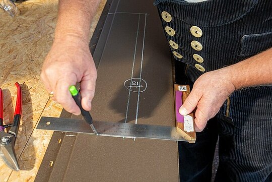 Detailaufnahme der PREFA Siding Paneele in Braun, auf der die Höhe der Ausklinkung, der Rückkantung sowie des Winkelschlages durch den Techniker angezeichnet werden.