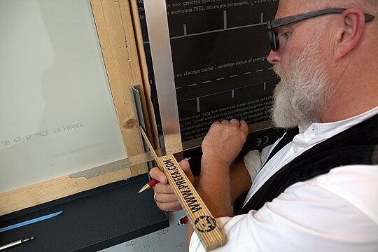 Techniker Tom Weiß nimmt mit einem Maßstab das Schenkelmaß eines F-Profils (Flutzprofils), dass er seitlich an einer Holz-Fensterlaibung angebracht hat. 