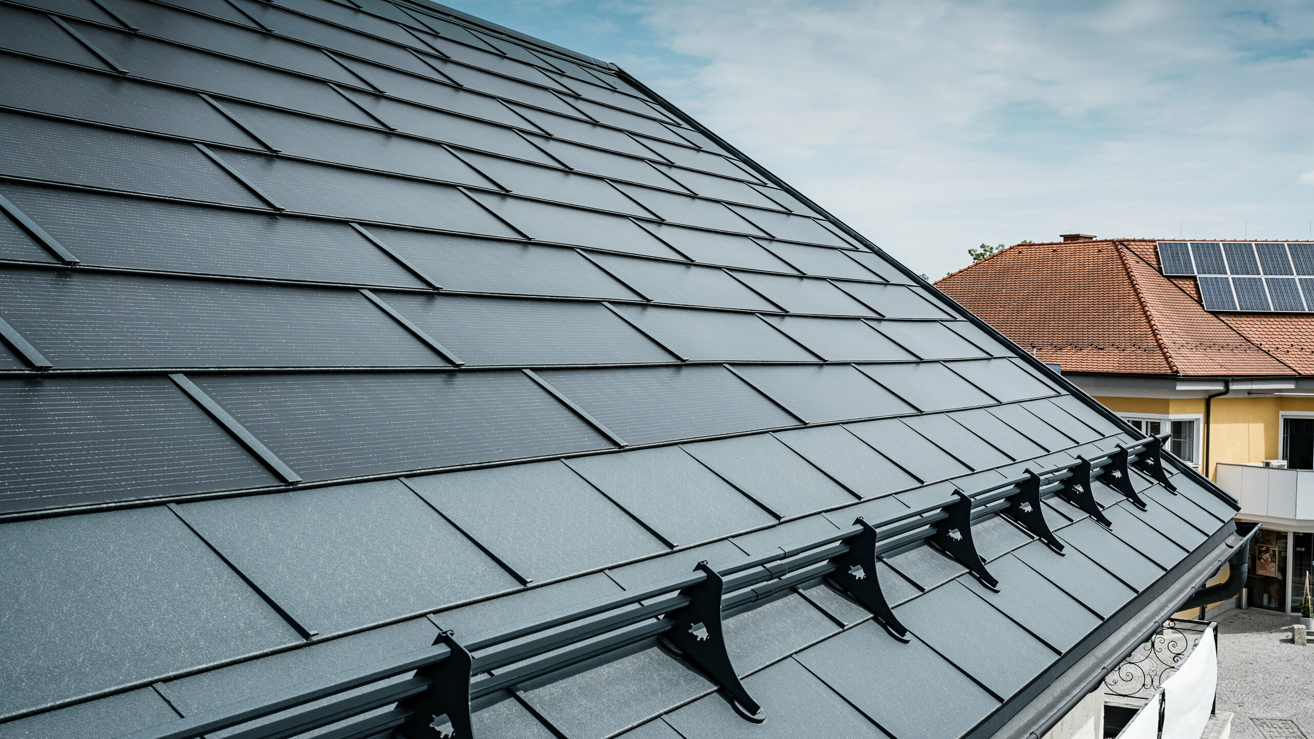 Ansicht eines modernen Daches mit anthrazitfarbenen PREFA Solardachplatten. In der Nähe der Traufe wurde das Dach mit klassischen PREFA Dachplatten R.16 und einem PREFA Schneeschutzsystem ausgestattet. Im Hintergrund ist das Dach eines benachbarten Hauses mit traditionellen Dachziegeln und Aufdach-Solarmodulen zu sehen, was den Kontrast zwischen neuer und herkömmlicher Dachgestaltung hervorhebt. Die innovative Solardachplatte von PREFA vereint Ästhetik und Funktionalität und bietet eine nachhaltige Energielösung für alle Gebäudearten.