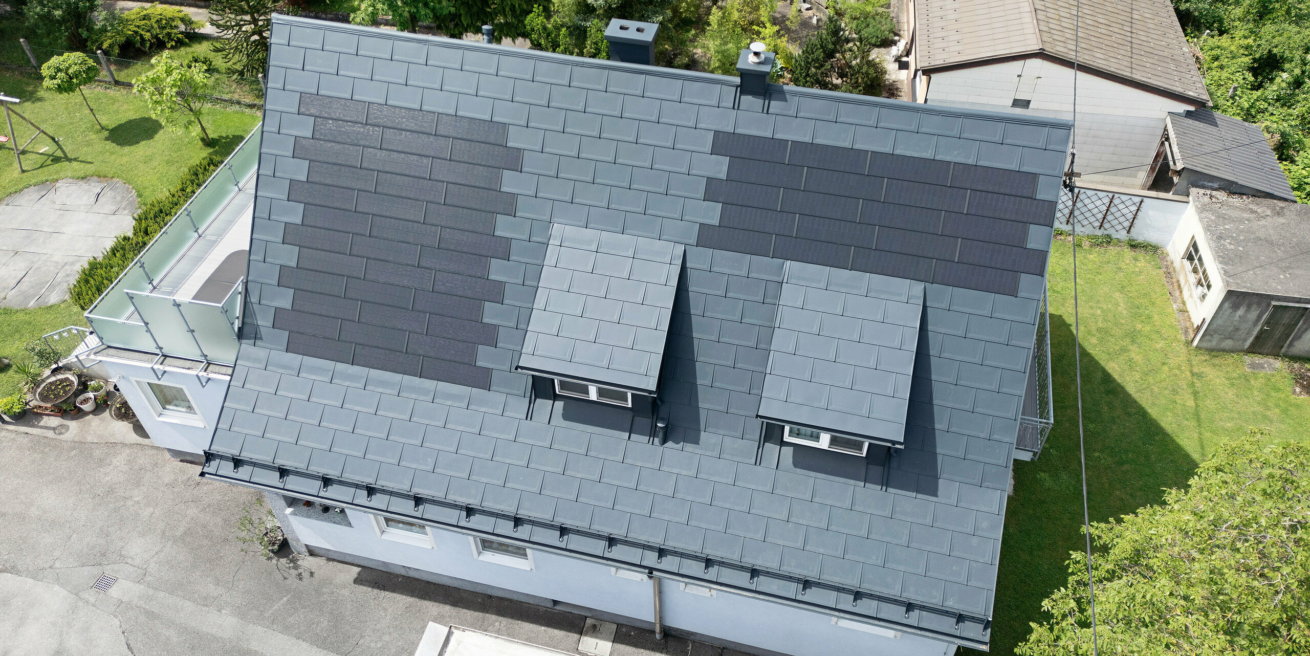 Komplett saniertes Dach eines Einfamilienhauses in Leonding mit PREFA Solardachplatten in Anthrazit. Die Vogelperspektive zeigt das modernisierte Dach mit integrierten Solarmodulen aus PREFA Aluminiumprodukten. Diese langlebige und witterungsbeständige Lösung bietet eine nachhaltige und energieeffiziente Möglichkeit zur Stromerzeugung. Das Bild verdeutlicht die ästhetische Integration der Solarmodule in die Dachfläche, die eine moderne und funktionale Architektur unterstützt. Die innovativen Solardachplatten wurden rund um zwei Schleppgauben platziert.