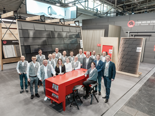 Gruppenfoto des PREFA Teams auf der BAU Messe vor dem Messestand, der innovative Lösungen für Dach, Fassade und Solar präsentiert. Im Hintergrund sind großformatige Fassaden- und Dachprodukte aus Aluminium sowie Informationswände zu sehen. Der Stand zeigt das Motto „Stark wie ein Stier“. Das Team trägt einheitliche, elegante Firmenkleidung.