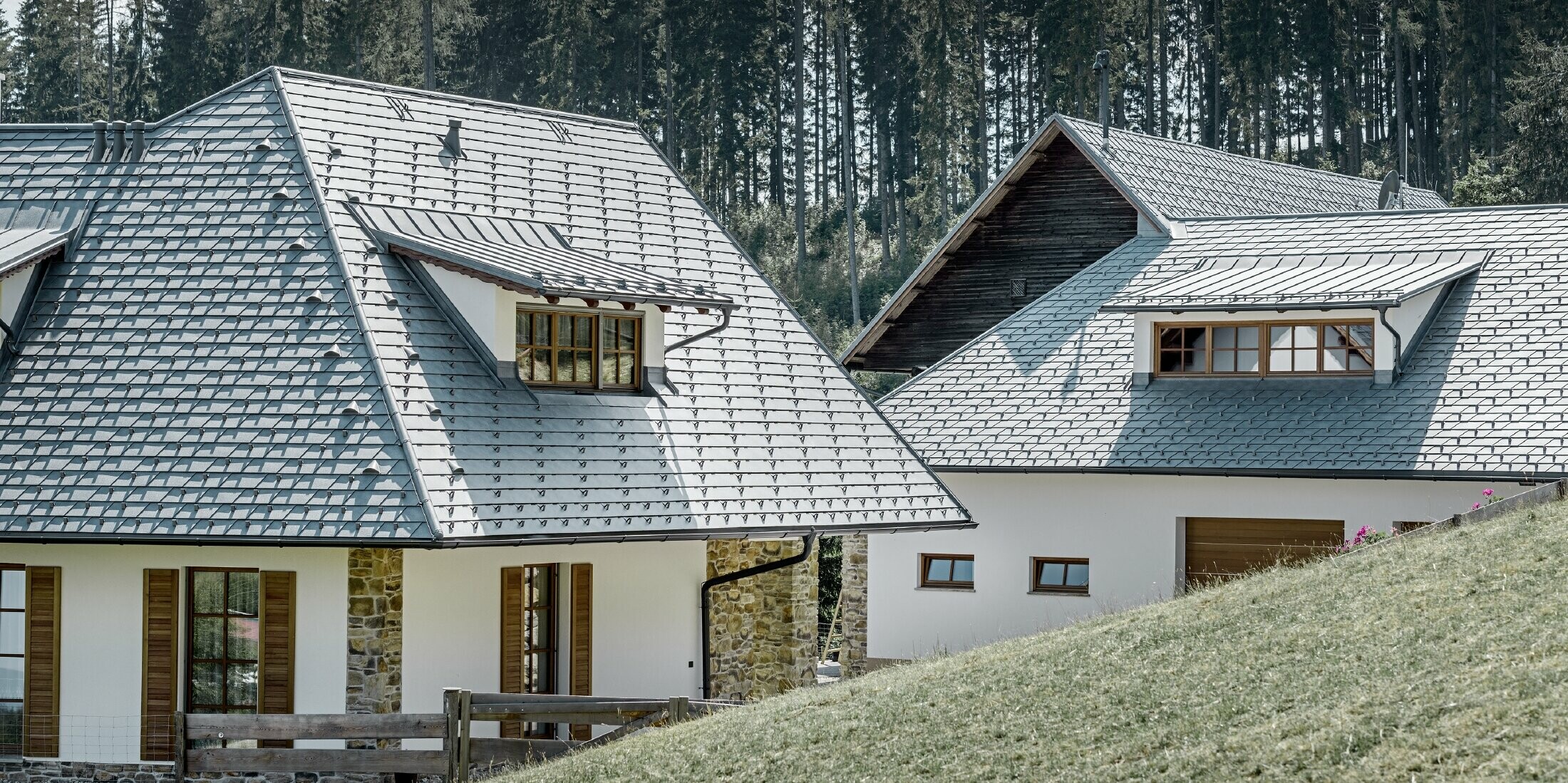 Rückansicht des Einfamilienhausees in Hanglage; Das Dach wurde mit der Alu-Dachschindel in Steingrau eingedeckt, die Schleppgauben mit Stehalz, ebenfalls in Steingrau. In Hintergrund erkennt man einen Wald.