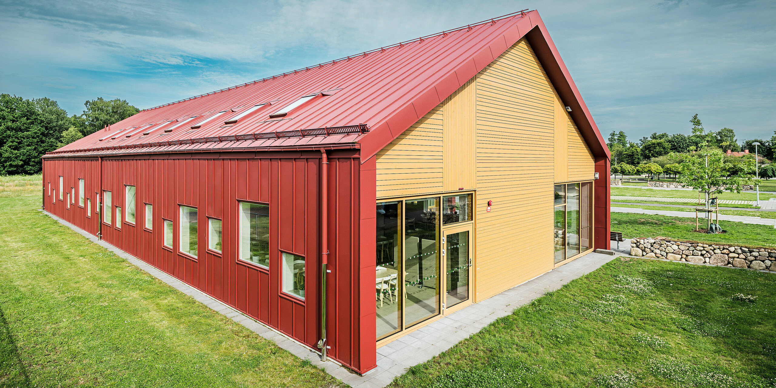 Die Giebelseite der Schulkantine Gröna Hjärtat in Broby, Schweden, zeigt eine beeindruckende Kombination aus PREFALZ in Oxydrot, Holz und großzügigen Glasflächen. Die Holzverkleidung an der Stirnseite bildet einen attraktiven Kontrast zur langlebigen Aluminiumverkleidung des restlichen Gebäudes. Mehrere Fenster und Dachfenster lassen viel natürliches Licht in das Gebäude, während das zeitgemäße Design die architektonische Qualität und Energieeffizienz des Bauwerks unterstreicht. Das Gebäude ist ein herausragendes Beispiel für zeitgenössische Architektur mit nachhaltigen Materialien und innovativer Gestaltung.