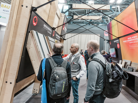 PREFA Außendienstler Fabio Fratello erklärt Messebesuchern am Messestand auf der BAU Messe die Konstruktion und Vorteile der PREFA Fassaden- und Dachelemente. Zu sehen sind verschiedene Musterexponate, die Aufbau und Materialeigenschaften veranschaulichen. Im Hintergrund wird das neue PREVARIO 3.S Solarmontagesystem präsentiert.