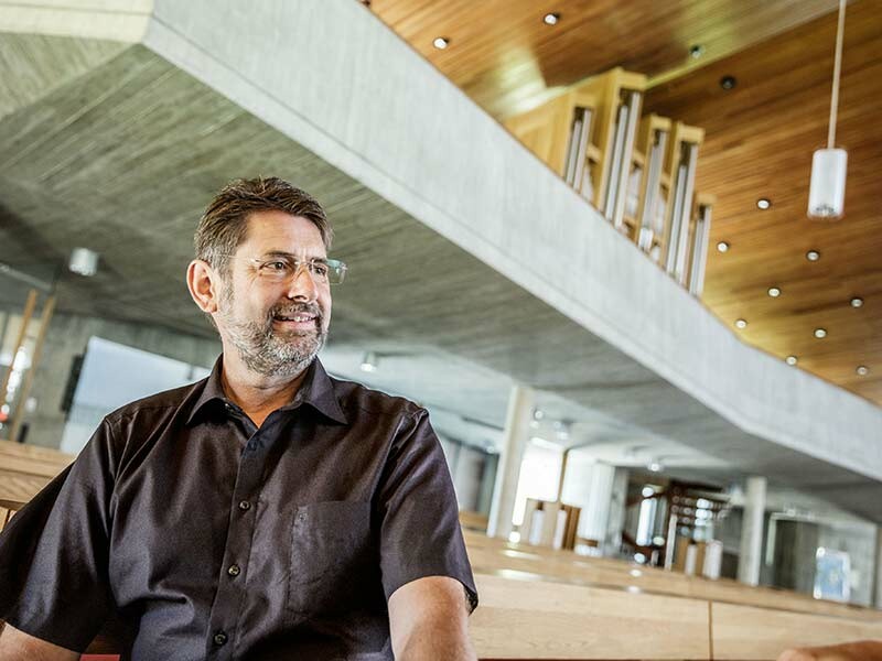 Portrait des Architekten Elmar Weber aus Langenau in der Kirche mit dunklem Hemd und Brille, mit einer Betonuntersicht der Galerie im Hintergrund und Holzbänken in der Kirche