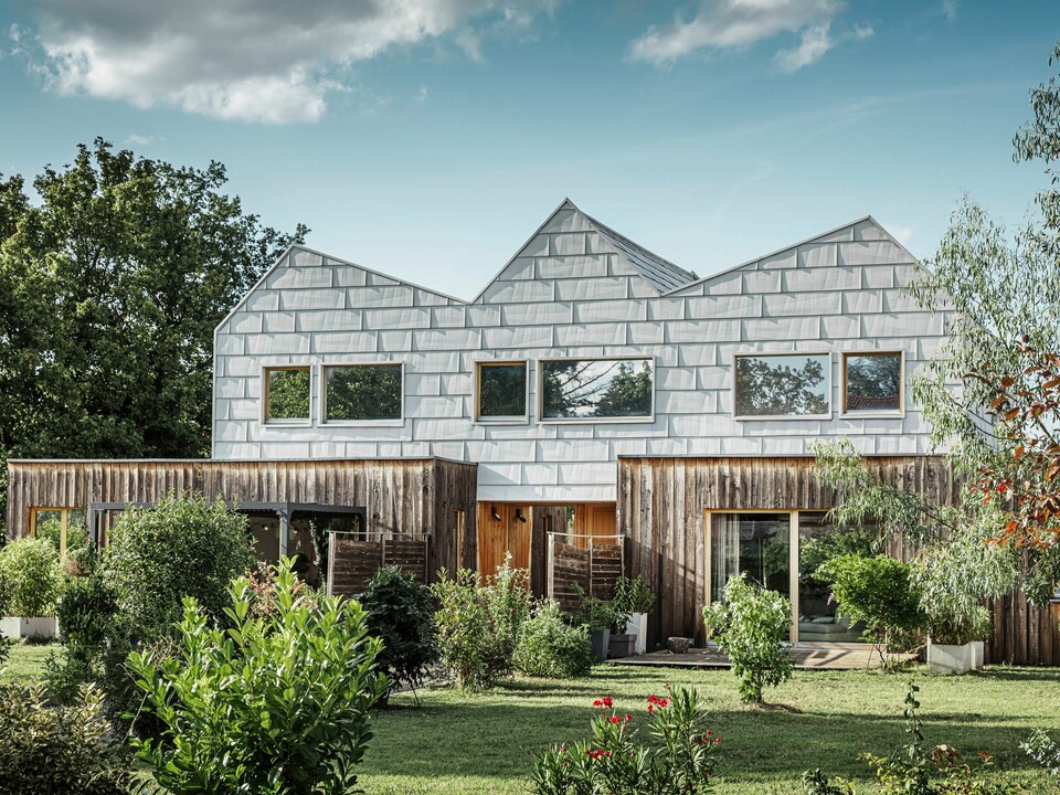 Modernes Doppelhaus mit Satteldach und PREFA-Fassade. Die Fassade besteht aus FX.12 Aluminiumpaneelen, die eine strukturierte, moderne Optik bieten. Das Haus ist von einem gepflegten Garten umgeben, und die Fensterrahmen harmonieren perfekt mit der Gesamtarchitektur.