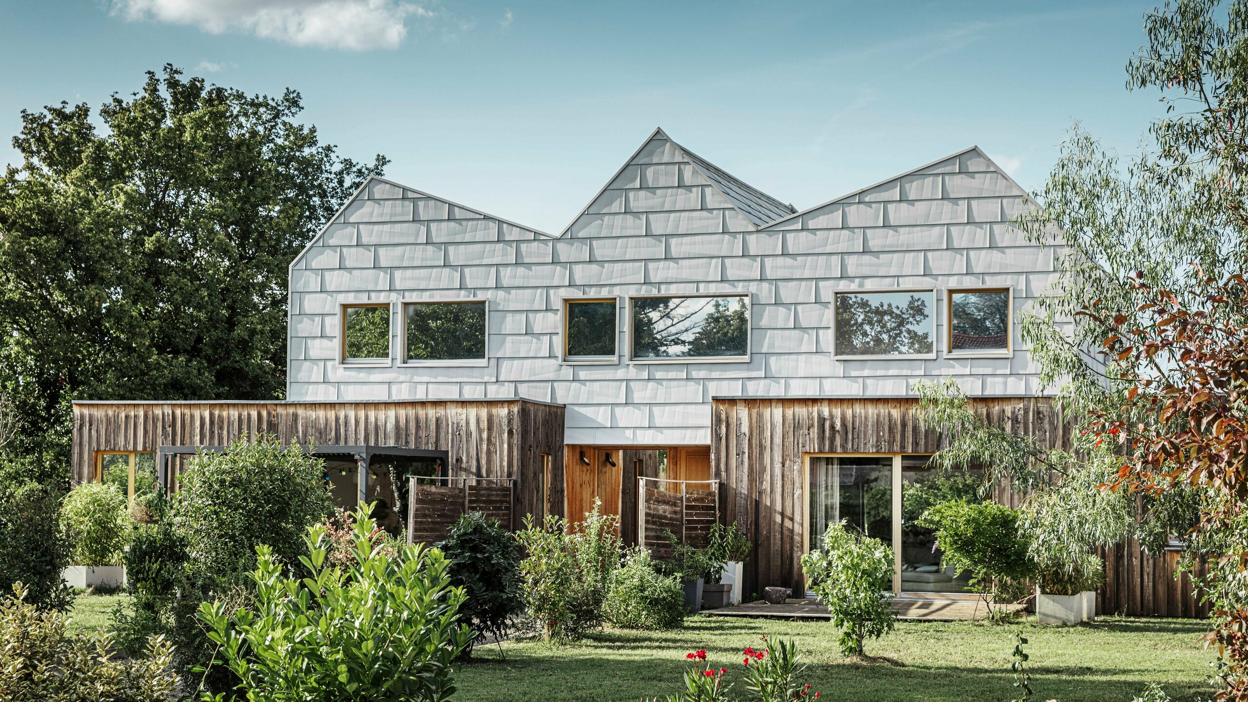 Modernes Doppelhaus mit Satteldach und PREFA-Fassade. Die Fassade besteht aus FX.12 Aluminiumpaneelen, die eine strukturierte, moderne Optik bieten. Das Haus ist von einem gepflegten Garten umgeben, und die Fensterrahmen harmonieren perfekt mit der Gesamtarchitektur.