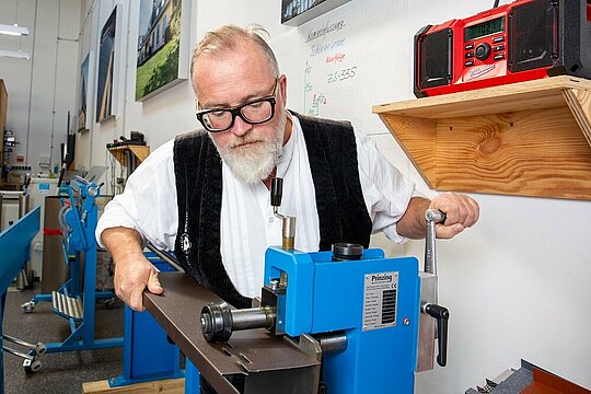 Auf diesem Bild ist neben dem PREFA Techniker eine blaue Maschine zu erkennen. Mithilfe dieser Maschine stanzt er die Zuluft-Löcher in das Sturzblech.