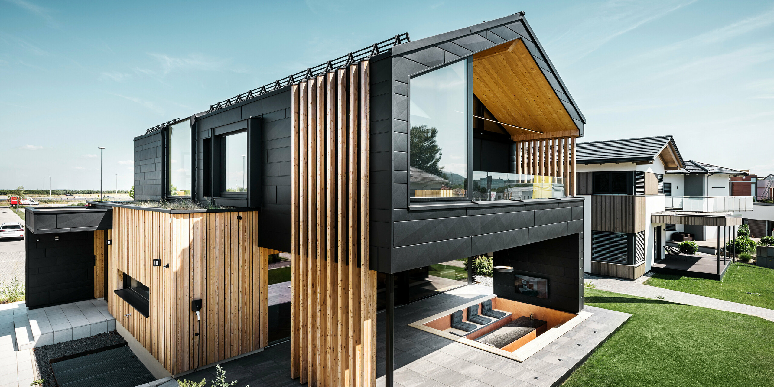 Seitenansicht des Musterhauses Autark in der Blauen Lagune in Vösendorf, Österreich. Das Obergeschoss besticht durch große Fensterflächen und eine moderne Aluminiumfassade aus PREFA Siding.X Paneelen in P.10 Schwarz, kombiniert mit vertikalen Holzelementen. Die großzügige Terrasse im Erdgeschoss mit eingelassener Sitzgelegenheit fügt sich nahtlos in die Architektur ein. Das stilvolle und nachhaltige Design des Hauses verbindet Aluminium und Holz auf eindrucksvolle Art und Weise.