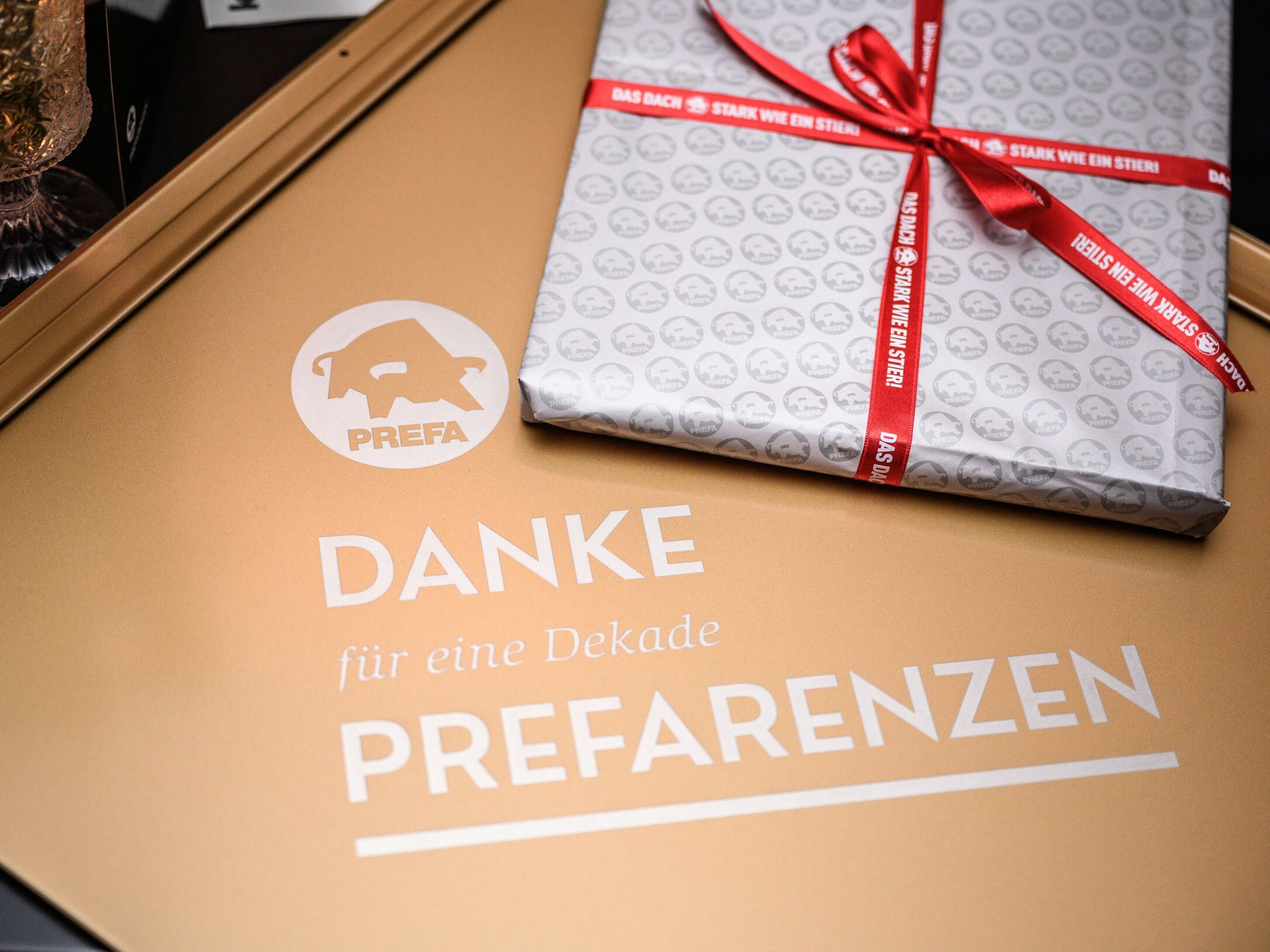 Ein Geschenk liegt auf einer goldenen Oberfläche mit dem PREFA Logo und dem Schriftzug 'DANKE für eine Dekade PREFARENZEN'. Das Geschenk ist in silbernes Papier eingepackt, das mit kleinen PREFA Logos bedruckt ist, und mit einem roten Band gebunden, auf dem 'STARK WIE EIN STIER' steht