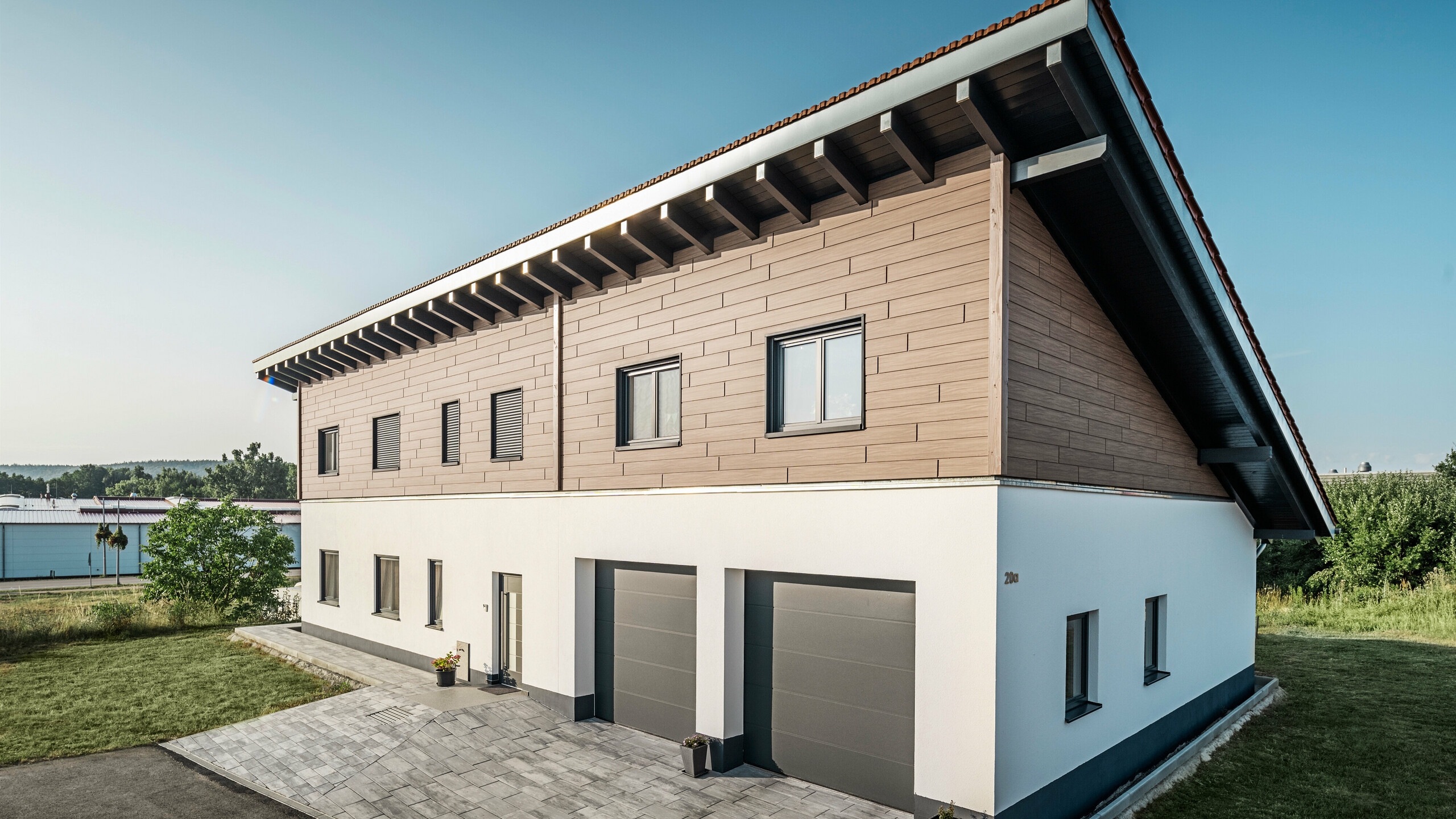 Blick von der Seite auf ein Einfamilienhaus mit Pultdach in Bruck. Das obere Stockwerk ist mit PREFA Sidings in Walnuss Braun ummantelt. Auf der rechten Seite des Bildes sind zwei Garagentore in Anthrazit zu sehen. In der Gebäudemitte befindet sich die Eingangstüre. Der Sockel ist in einem schlichten Weiß gehalten. 