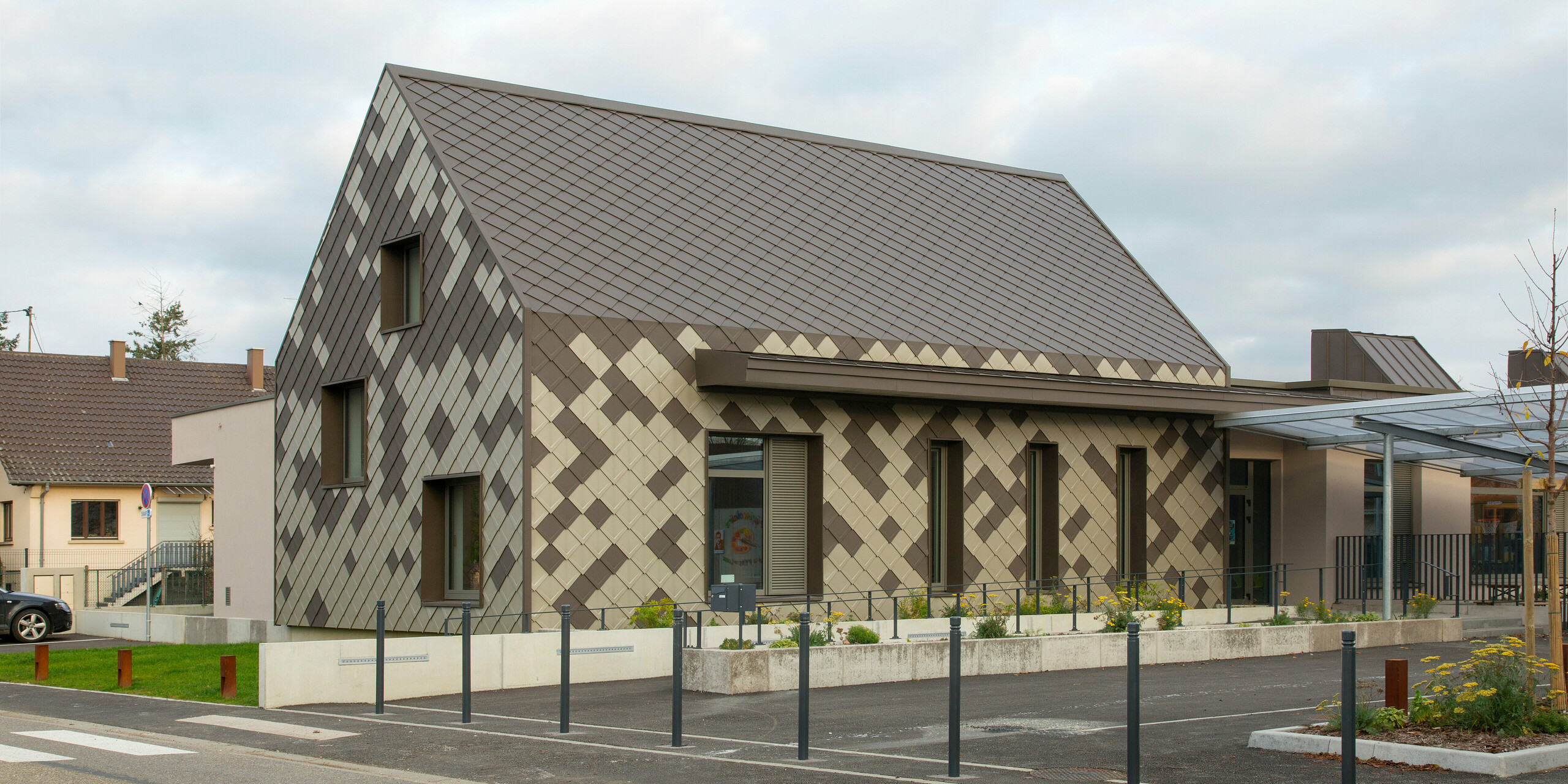 Schräger Blick auf den Hort in Sessenheim. Das Gebäude besitzt eine einprägsame Gebäudehülle aus Aluminium in den Farben Braun und Bronze.