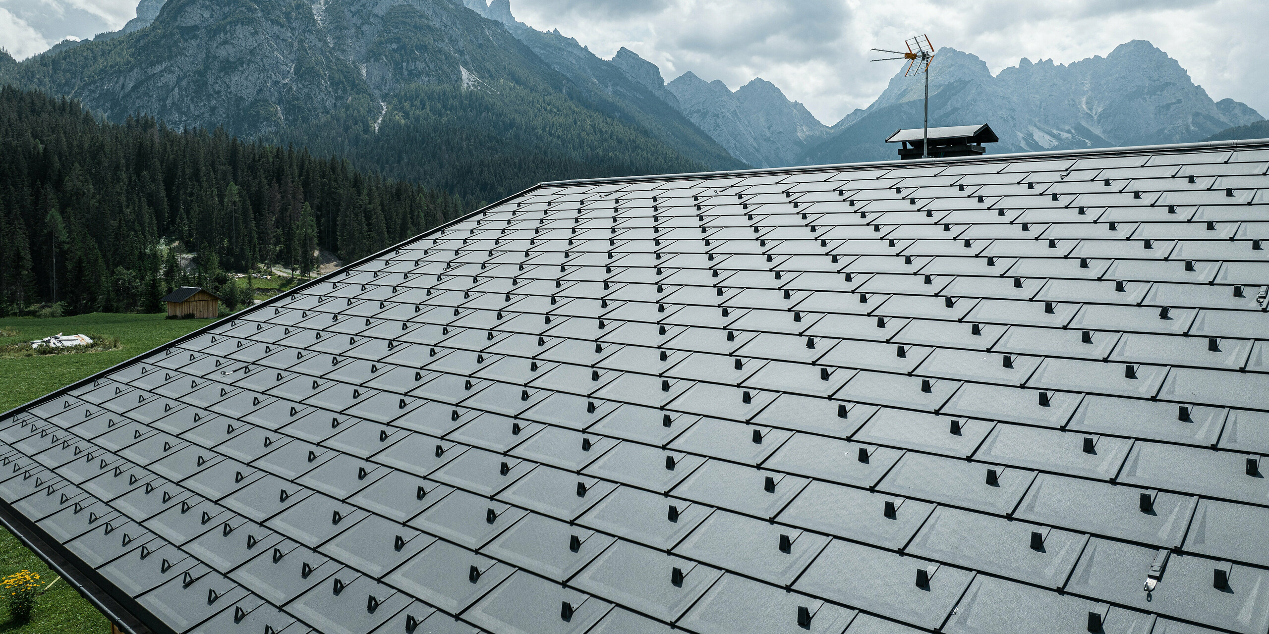 Casa Hoffe in Sappada, Italien, geprägt durch die PREFA Dachplatten R.16 in P.10 Schwarz. Das Dachsystem fügt sich hervorragend in die natürliche Umgebung der Dolomiten ein. Die präzise verlegten Dachplatten bieten eine effiziente und dauerhafte Bedeckung, die das traditionelle Erscheinungsbild des Hauses mit modernster Technik und Materialbeständigkeit vereint. Der Anblick des robusten Aluminiumdachs vor dem Hintergrund der beeindruckenden Bergkulisse symbolisiert die Verschmelzung von Natur und fortschrittlicher Bautechnologie.