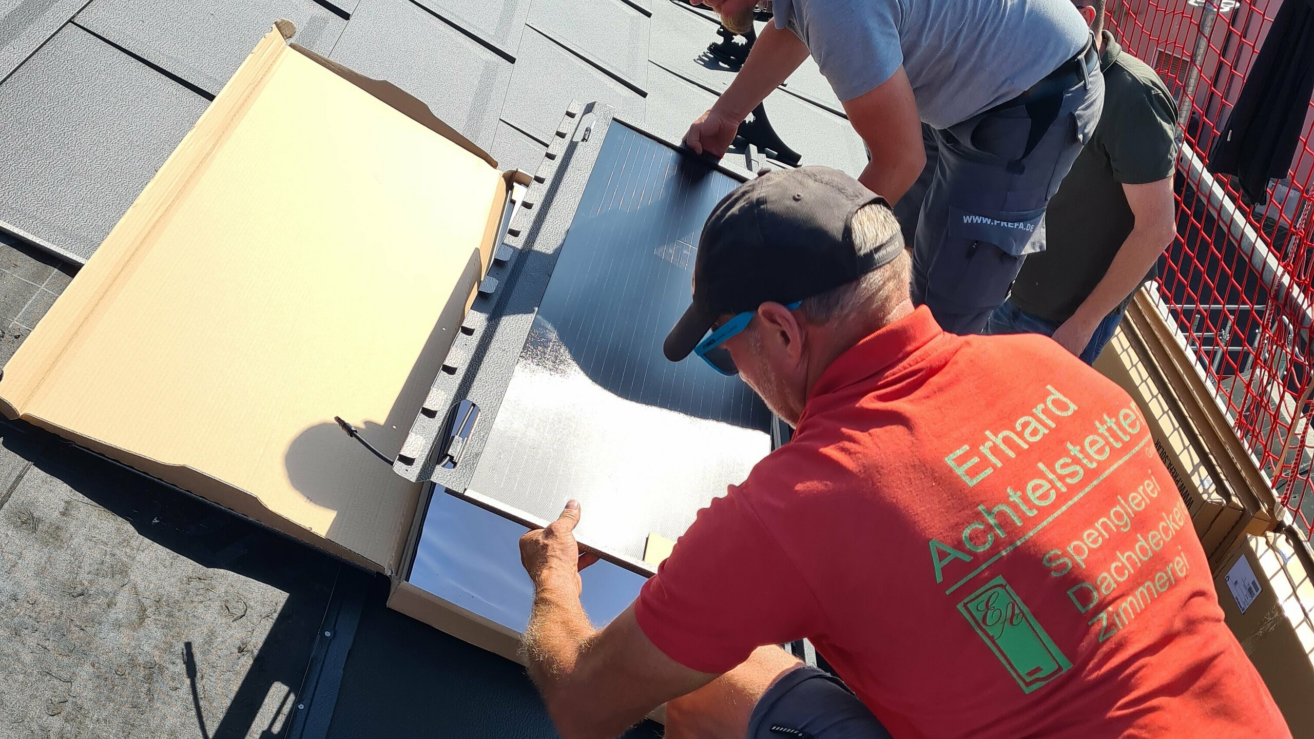 Ein Karton mit PREFA-Solardachplatten wurde auf dem Dach der Kirche geöffnet. Einige Solardachplatten sind sichtbar. Die Platten sind ordentlich gestapelt und haben eine schwarze, glänzende Oberfläche. Auf dem Karton ist das PREFA-Logo und weitere Informationen zum Produkt abgedruckt.