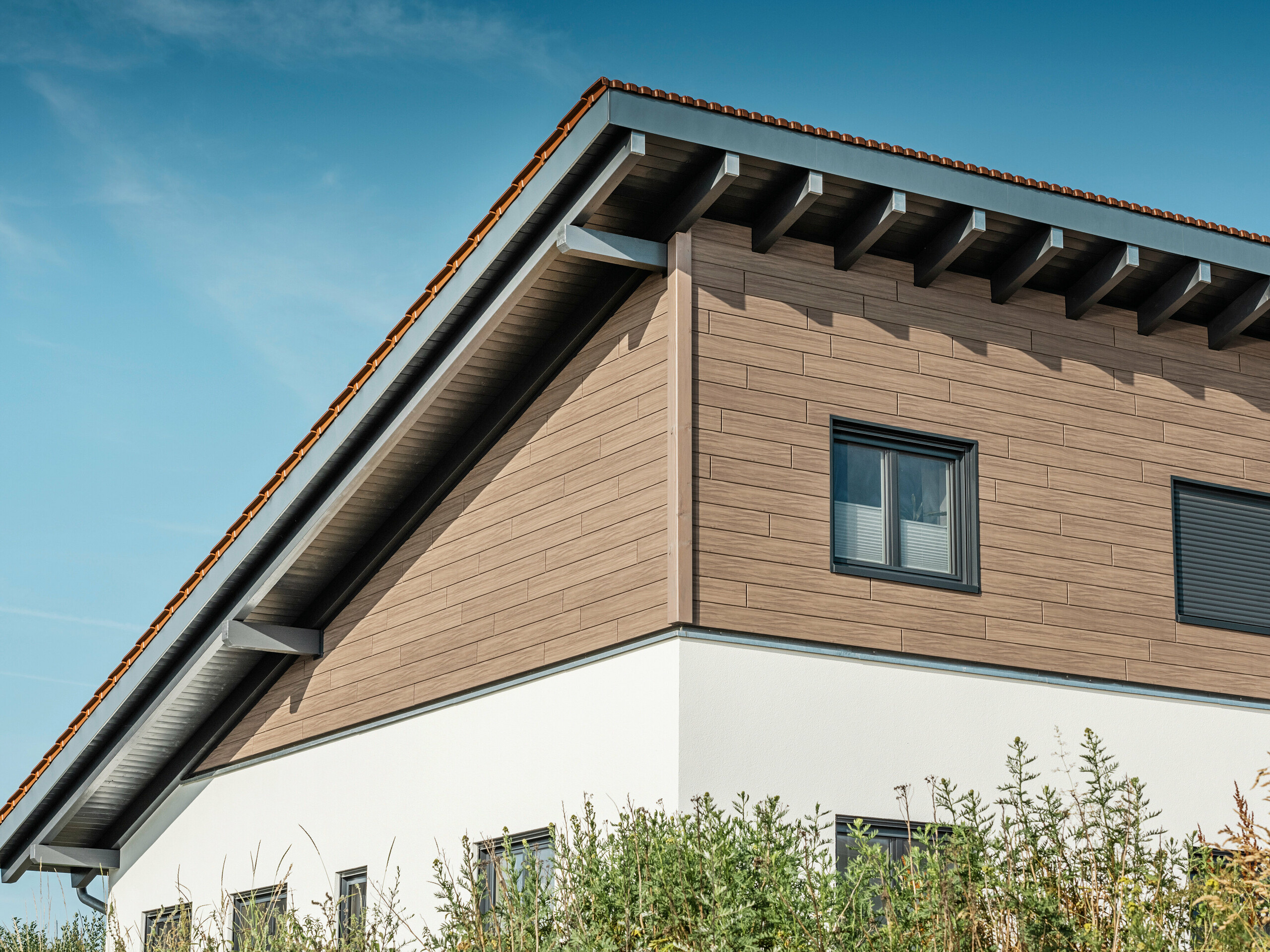 Schräger Blick von unten auf das Obergeschoss eines Einfamilienhauses in Bruck. Das obere Stockwerk ist mit PREFA Sidings in einer Holzoptik umhüllt. Zu sehen ist das Pultdach, das auf der Gebäuderückseite nach unten verläuft.