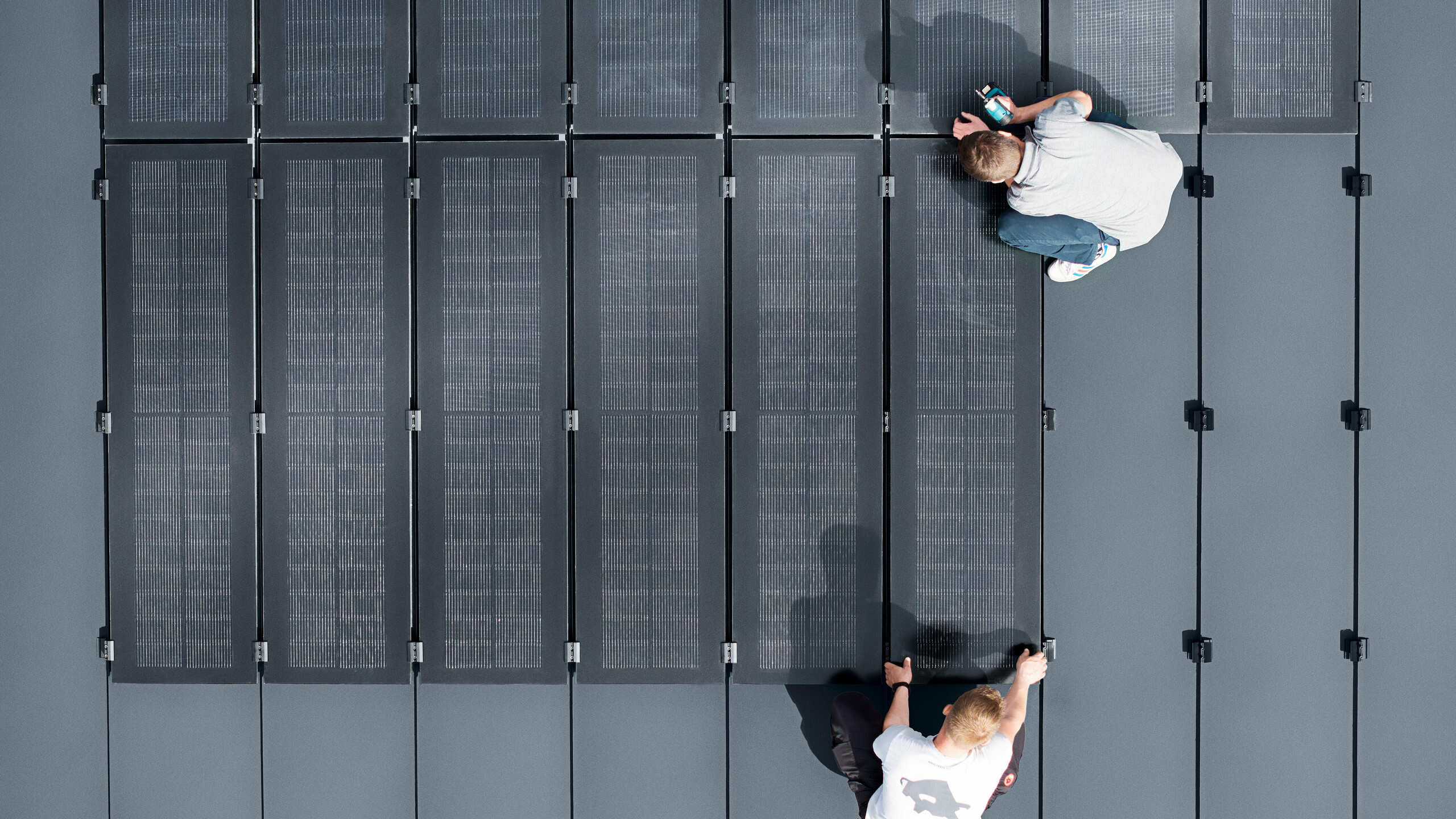 Zwei Personen installieren PREFALZ Solarmodule auf einer großen grauen Dachfläche, die mit Doppelstehfalzbahnen ausgelegt ist.