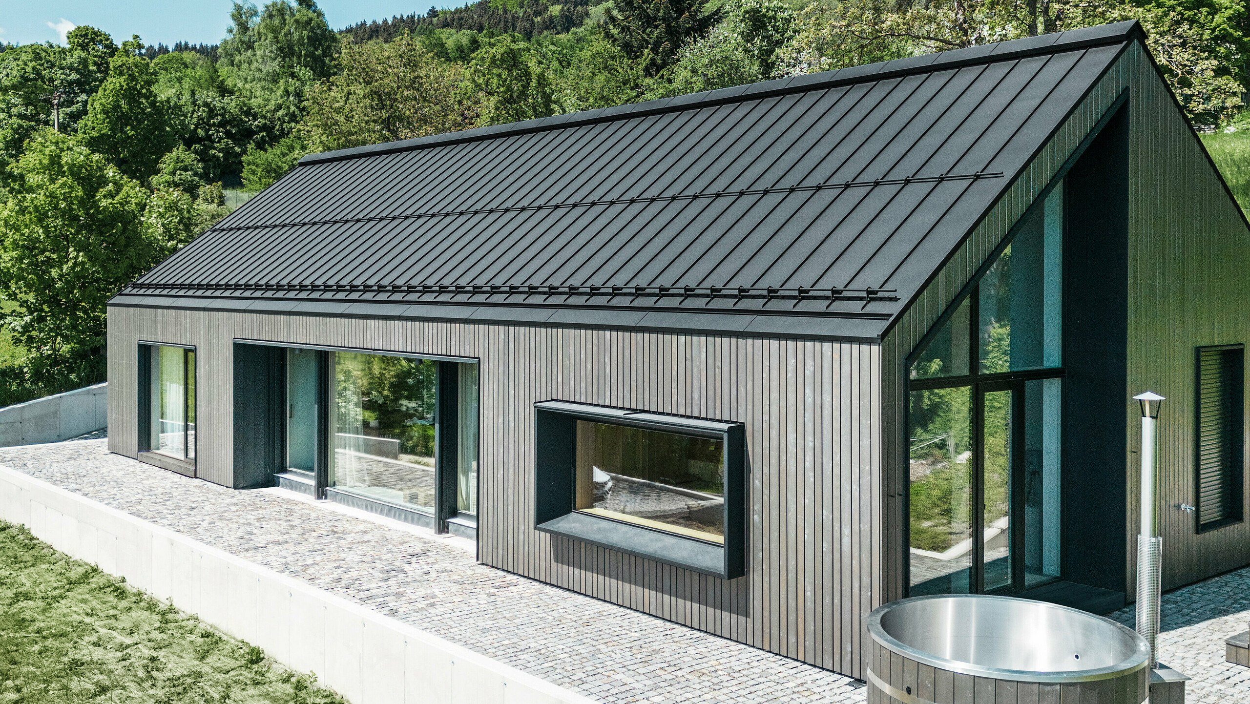 Ein modernes Holzmassivhaus in Kuncice (CZ) wurde mit großen Fensterflächen ausgestattet. Das dunkle Holzmassivhaus verfügt über ein Dachsystem aus PREFALZ in der Farbe P. 10 Schwarz. Im hinteren Außenbereich steht ein Whirlpool mit einer Holzverkleidung.