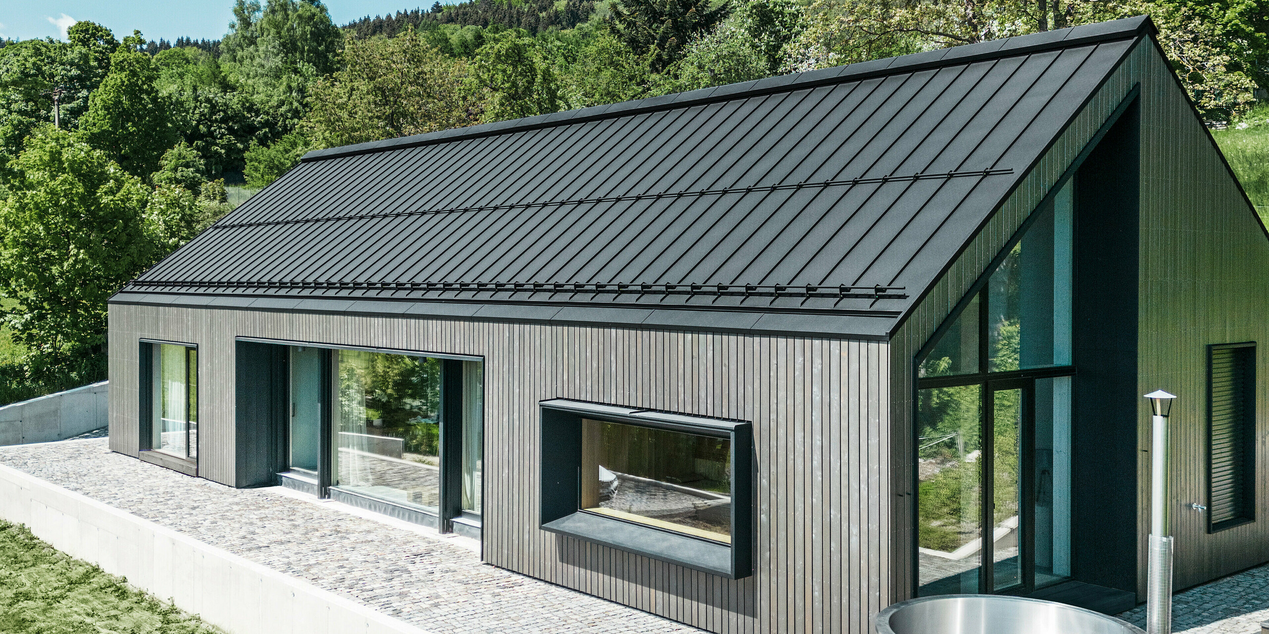 Ein modernes Holzmassivhaus in Kuncice (CZ) wurde mit großen Fensterflächen ausgestattet. Das dunkle Holzmassivhaus verfügt über ein Dachsystem aus PREFALZ in der Farbe P. 10 Schwarz. Im hinteren Außenbereich steht ein Whirlpool mit einer Holzverkleidung.