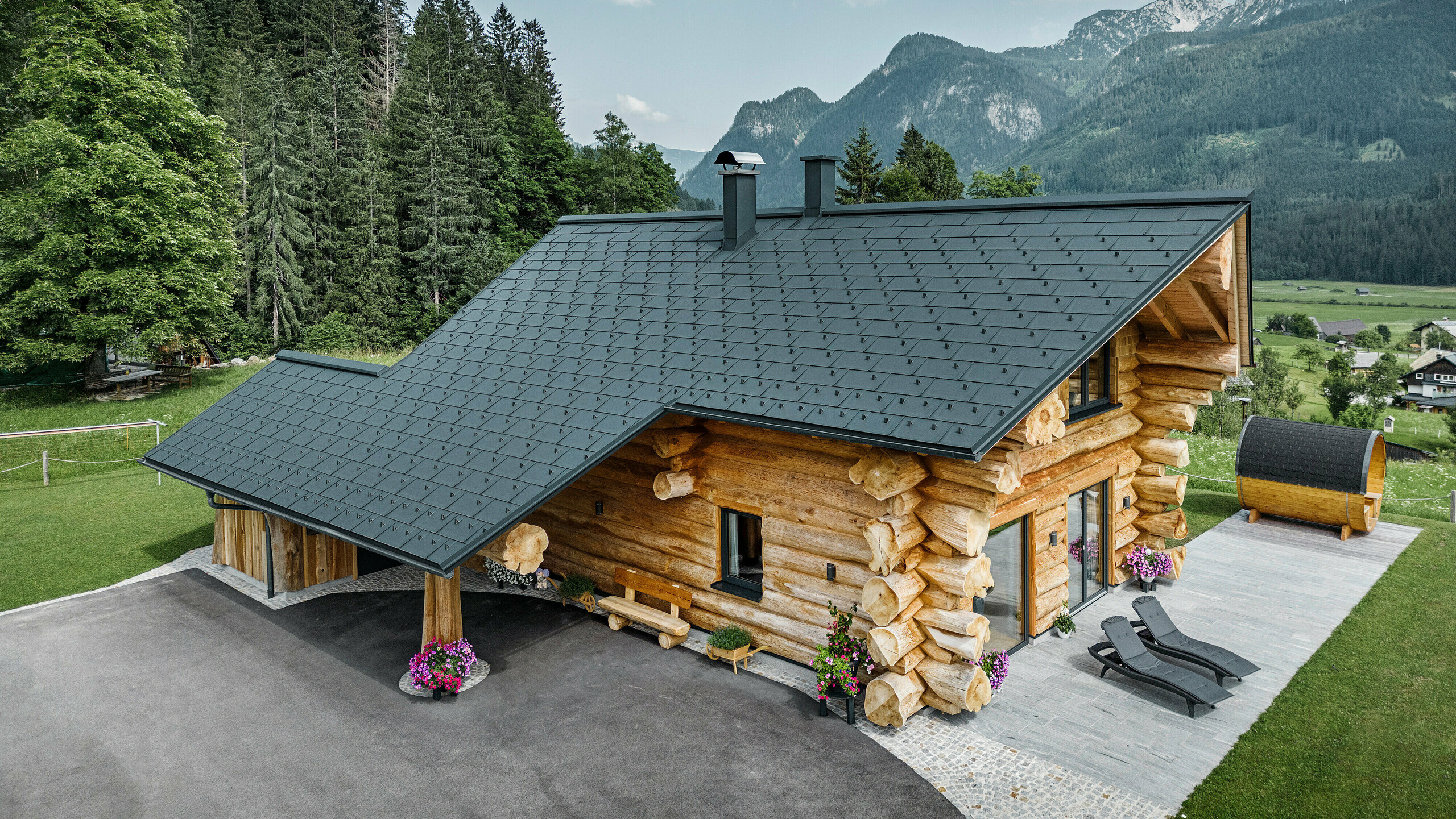 Schräge Ansicht eines malerischen Blockhauses in Gosau, Österreich, ausgestattet mit einem PREFA Dach aus anthrazitfarbenen Dachplatten R.16. Die Architektur kombiniert rustikales Blockhausflair mit modernen Elementen, umgeben von dichten grünen Wäldern und einer beeindruckenden Bergkulisse. Das Haus strahlt mit seiner ländlichen Eleganz und den präzise verlegten Dachelementen aus Aluminium, die für ihre Langlebigkeit und Witterungsbeständigkeit bekannt sind, eine einladende Atmosphäre aus.