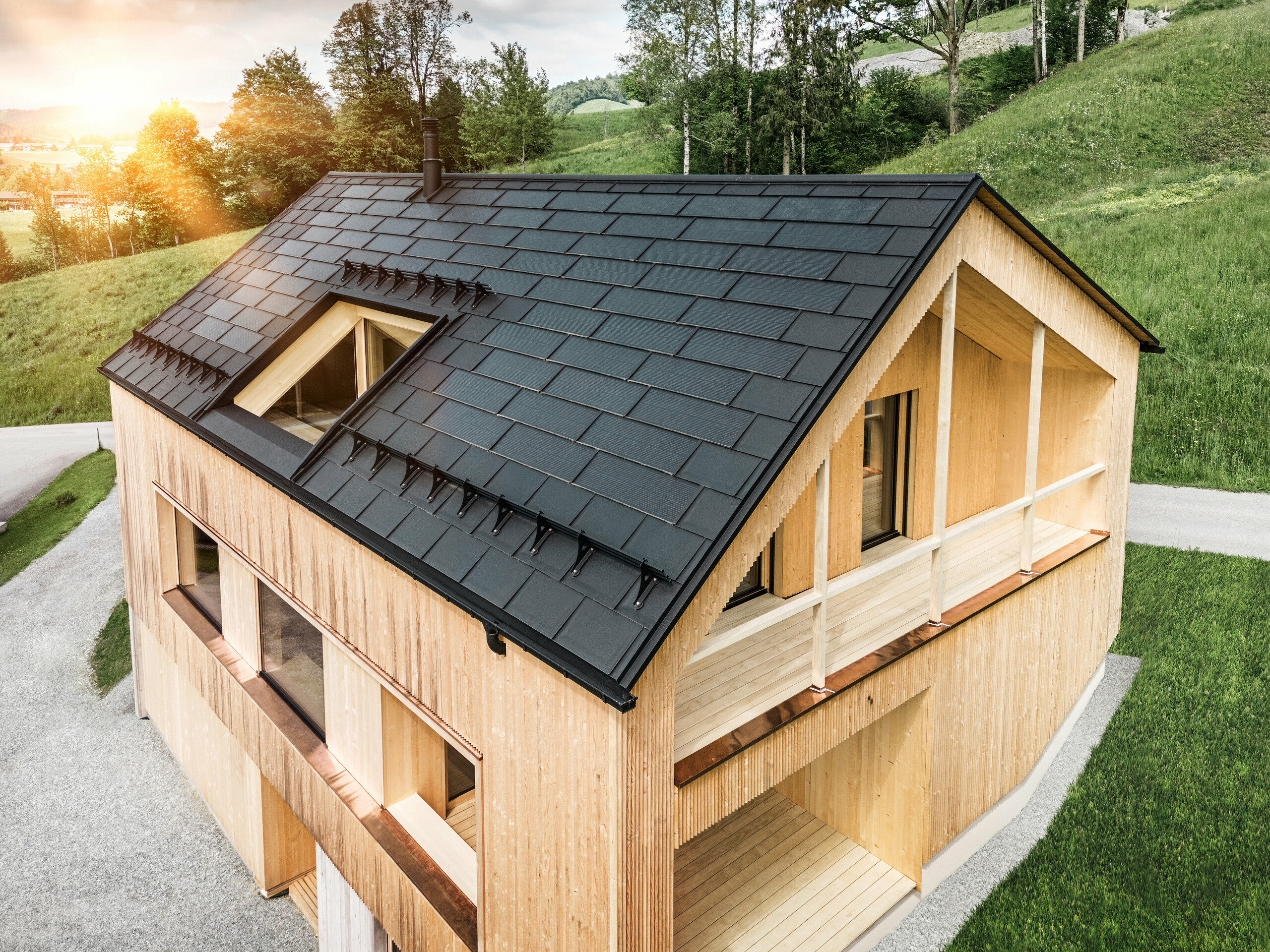 Blick auf ein modernes, nachhaltiges Holzhaus in Egg mit einem PREFA Solardach, das durch seine dunkle, strukturierte sowie elegante Erscheinung auffällt. Die Solarmodule sind strategisch zwischen den klassischen Dachplatten R.16 platziert, um eine optimale Sonneneinstrahlung zu nutzen und sich dabei ästhetisch in das Gesamtdesign einzufügen. Das Haus steht im Einklang mit seiner grünen Umgebung, während die warme Abendsonne die natürliche Holztextur der Fassade unterstreicht und die solide Bauweise betont. Ein harmonisches Beispiel für umweltbewusstes Bauen und Energieeffizienz.