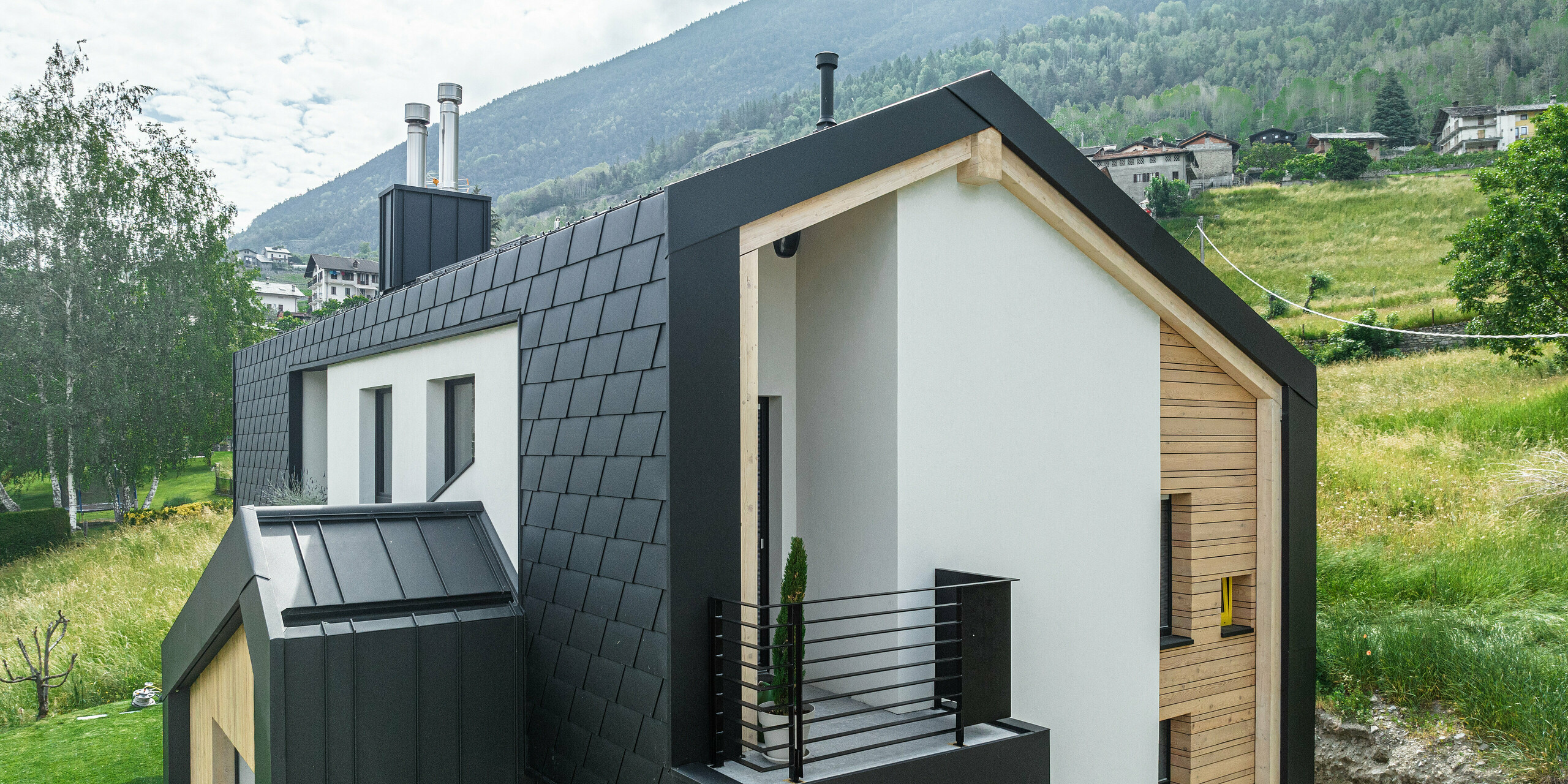 Moderne Architektur trifft auf Funktionalität bei diesem Einfamilienhaus, das von PREFA Dachschindeln DS.19 und Wandschindeln in Schwarz umhüllt ist. Der Blickwinkel betont die saubere Linienführung und die Kombination aus dunklen Schindeln und natürlichen Holzelementen, während das Haus harmonisch in die bergige Landschaft integriert ist.