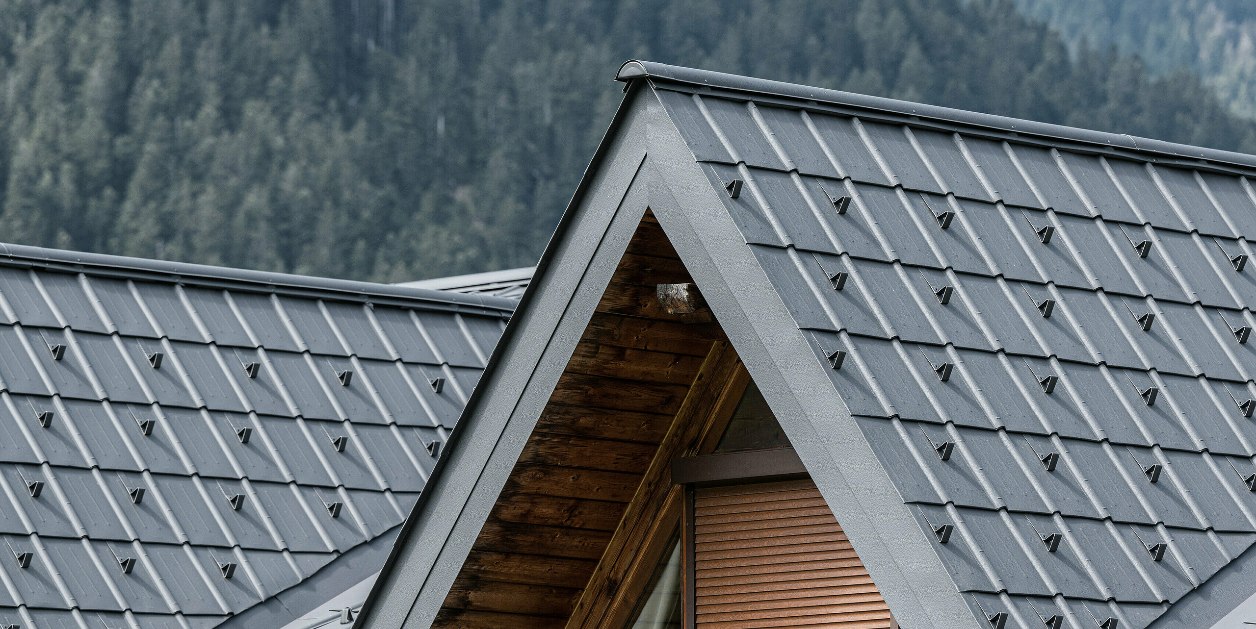 Detailní záběr na vikýř na alpském domě v Auronzo di Cadore, vybavený falcovanými taškami PREFA ve světle šedé barvě. Jemně strukturované hliníkové panely krásně kontrastují s přírodním dřevem střešní konstrukce a nabízejí vysoce kvalitní ochranu před alpským počasím a zároveň se hladce začleňují do horské lesní krajiny.