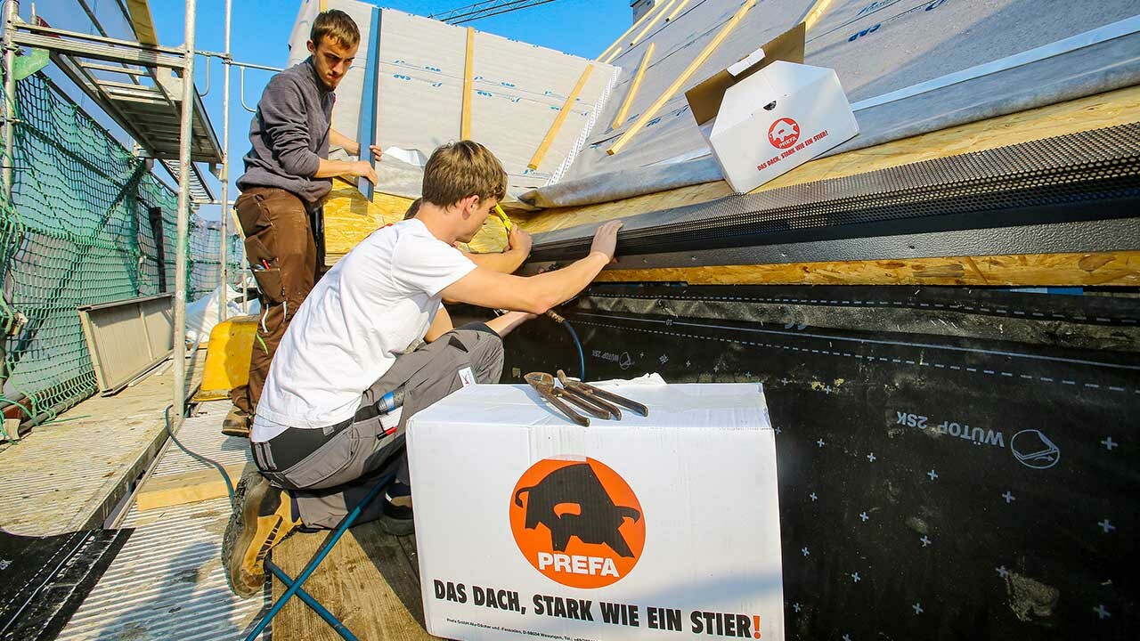 die Klempner der Firma Schütz bei der Arbeit, sie montieren den PREFA Saumstreifen für die Dachschindel, im Vordergrund steht ein PREFA Karton