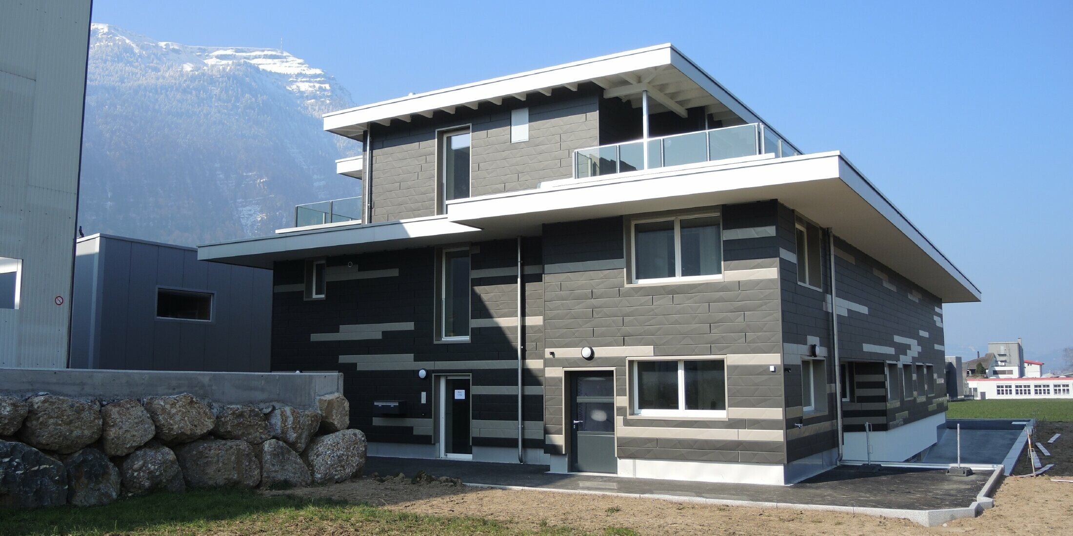 Individuelle Fassadengestaltung mit der vorgehängten hinterlüfteten Alufassade von PREFA. Die Fassade wurde mit den Fassadenpaneelen Siding.X in den Farben schwarzgrau und bronze gestaltet. Die Paneele haben eine geknickte Optik und wirf somit ein interessantes Schattenmuster.