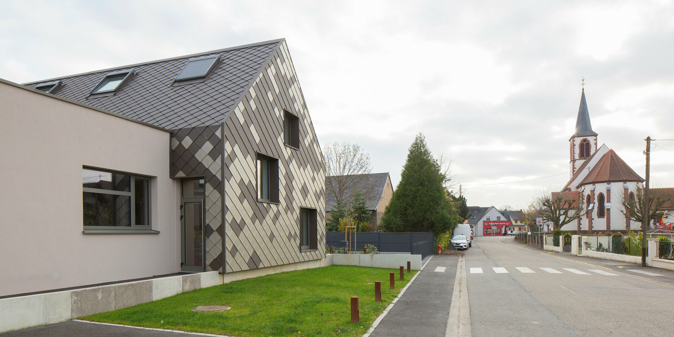 Eine Straße in Sessenheim: Auf der linken Seite der Hort mit der anschaulichen Aluminiumhülle, auf der rechten Seite eine Kirche.