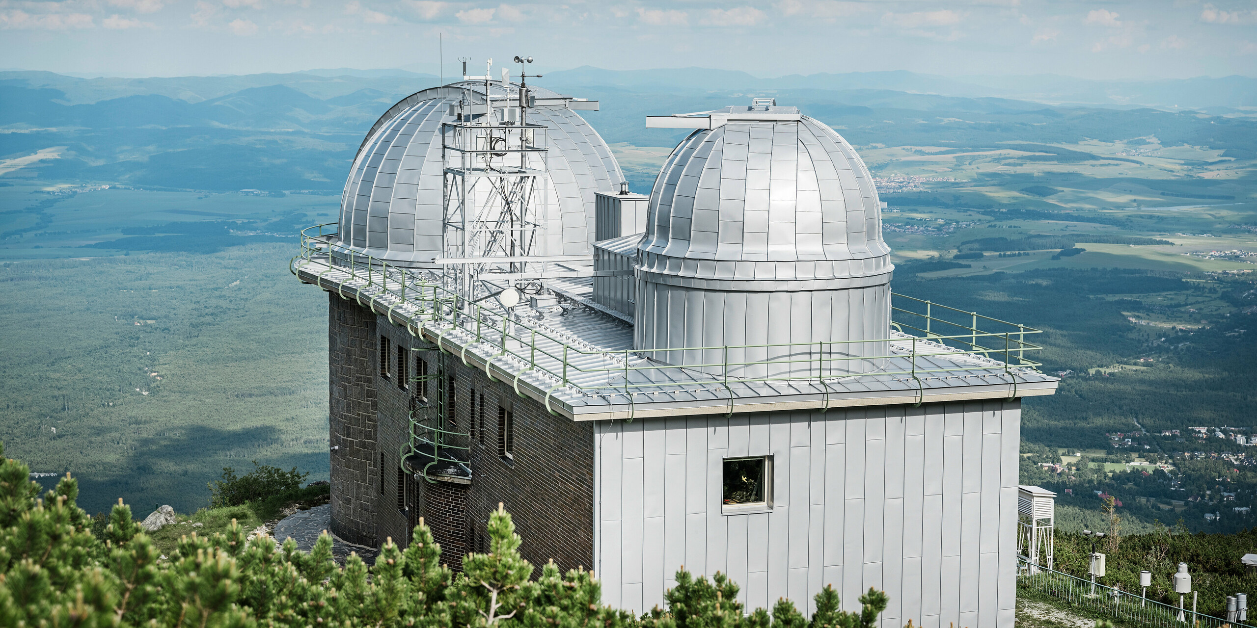 Rückansicht der Sternwarte Skalnaté Pleso, kunstvoll gekrönt mit dem PREFALZ Dach- und Fassadensystem in Silbermetallic. Im Vordergrund die robuste Aluminiumverkleidung, im Hintergrund ein atemberaubendes Panorama der slowakischen Tiefebene. Dieses Bauwerk ist ein Paradebeispiel für die Integration von modernen Materialien und traditioneller Architektur.