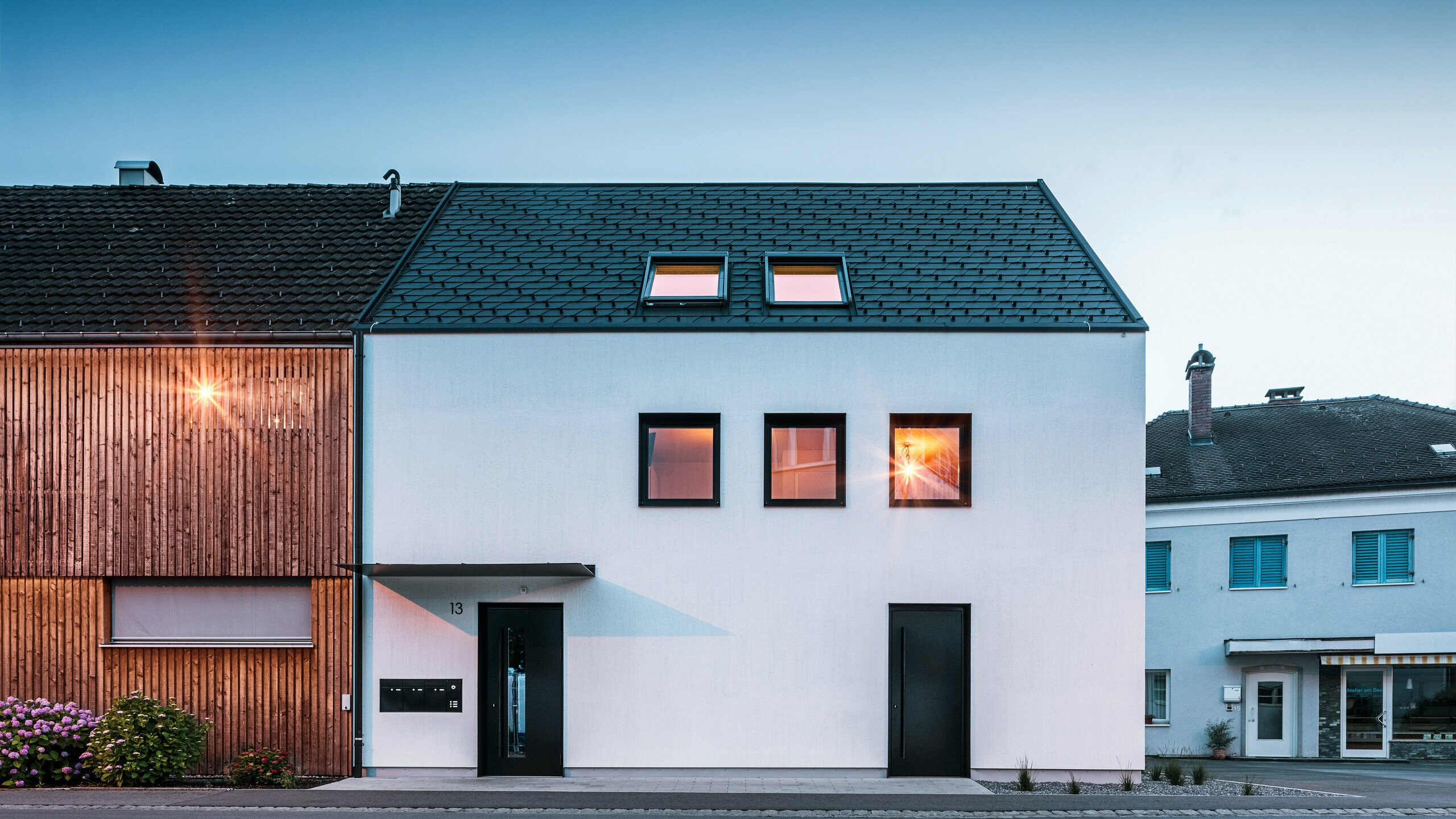 Moderne Mehrfamilienhausfassade mit PREFA Dachschindeln DS19 und Prefalz in Schwarz am Bodensee in Österreich. Das Gebäude hat eine klare Zweiteilung mit einem hölzernen Anbau auf der linken Seite und einem verputzten Hauptteil auf der rechten Seite. Das Dach zeigt die charakteristische dunkle Schindeldeckung und es gibt mehrere rechteckige Fenster, die im warmen Abendlicht spiegeln.