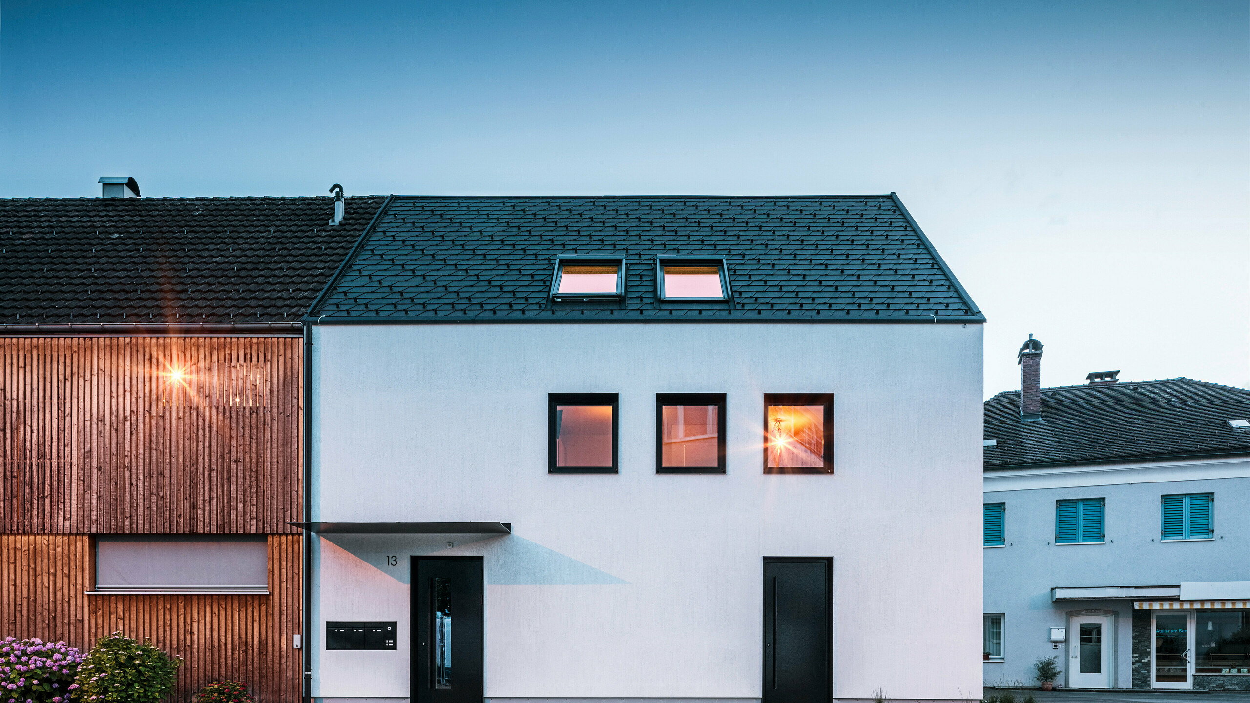 Moderne Mehrfamilienhausfassade mit PREFA Dachschindeln DS19 und Prefalz in Schwarz am Bodensee in Österreich. Das Gebäude hat eine klare Zweiteilung mit einem hölzernen Anbau auf der linken Seite und einem verputzten Hauptteil auf der rechten Seite. Das Dach zeigt die charakteristische dunkle Schindeldeckung und es gibt mehrere rechteckige Fenster, die im warmen Abendlicht spiegeln.