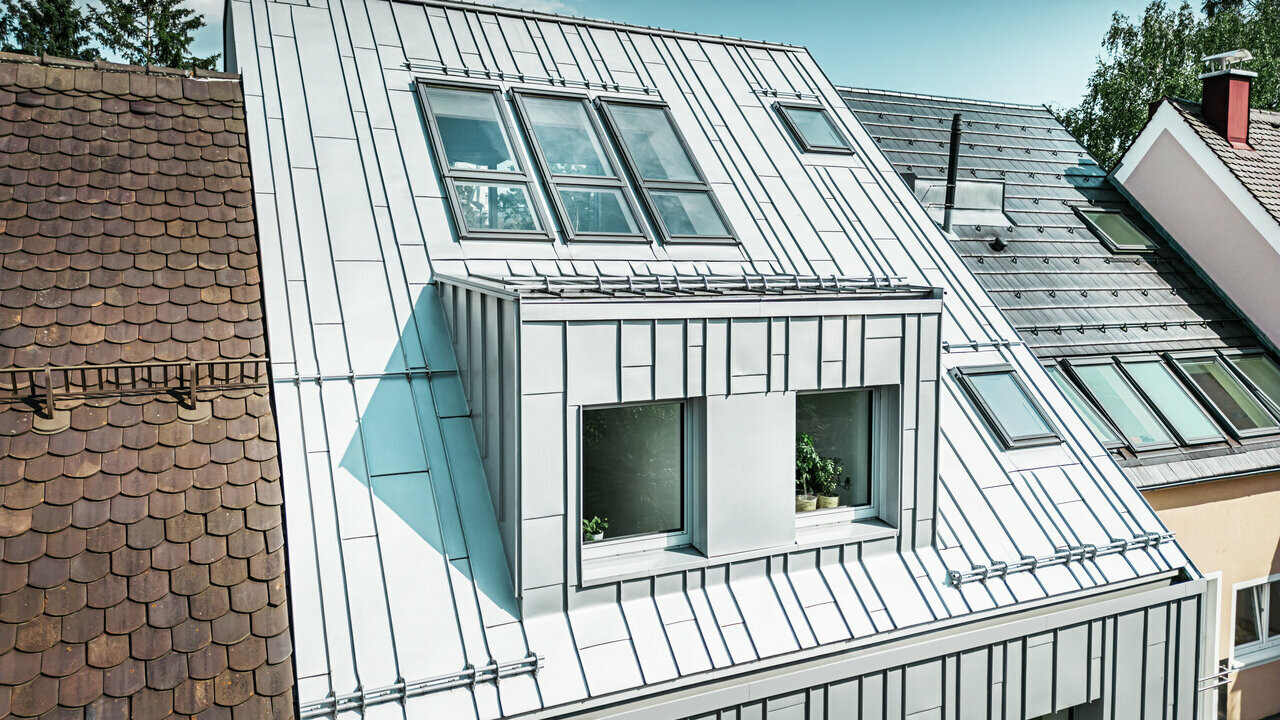 Ein modernes Reihenhaus mit einer Dach- und Fassadensanierung, die PREFA Aluminiumprodukte zeigt. Das Dach besteht aus silbermetallischen, kleinformatigen, stehfalzartigen Aluminiumplatten, die eine strukturierte, matte Oberfläche aufweisen und das Licht reflektieren. Die Hausfassade ist ebenfalls mit vertikalen Aluminiumelementen verkleidet, die einen zeitgemäßen Kontrast zu den traditionellen, terrakottafarbenen Ziegeldächern der angrenzenden Häuser bieten.