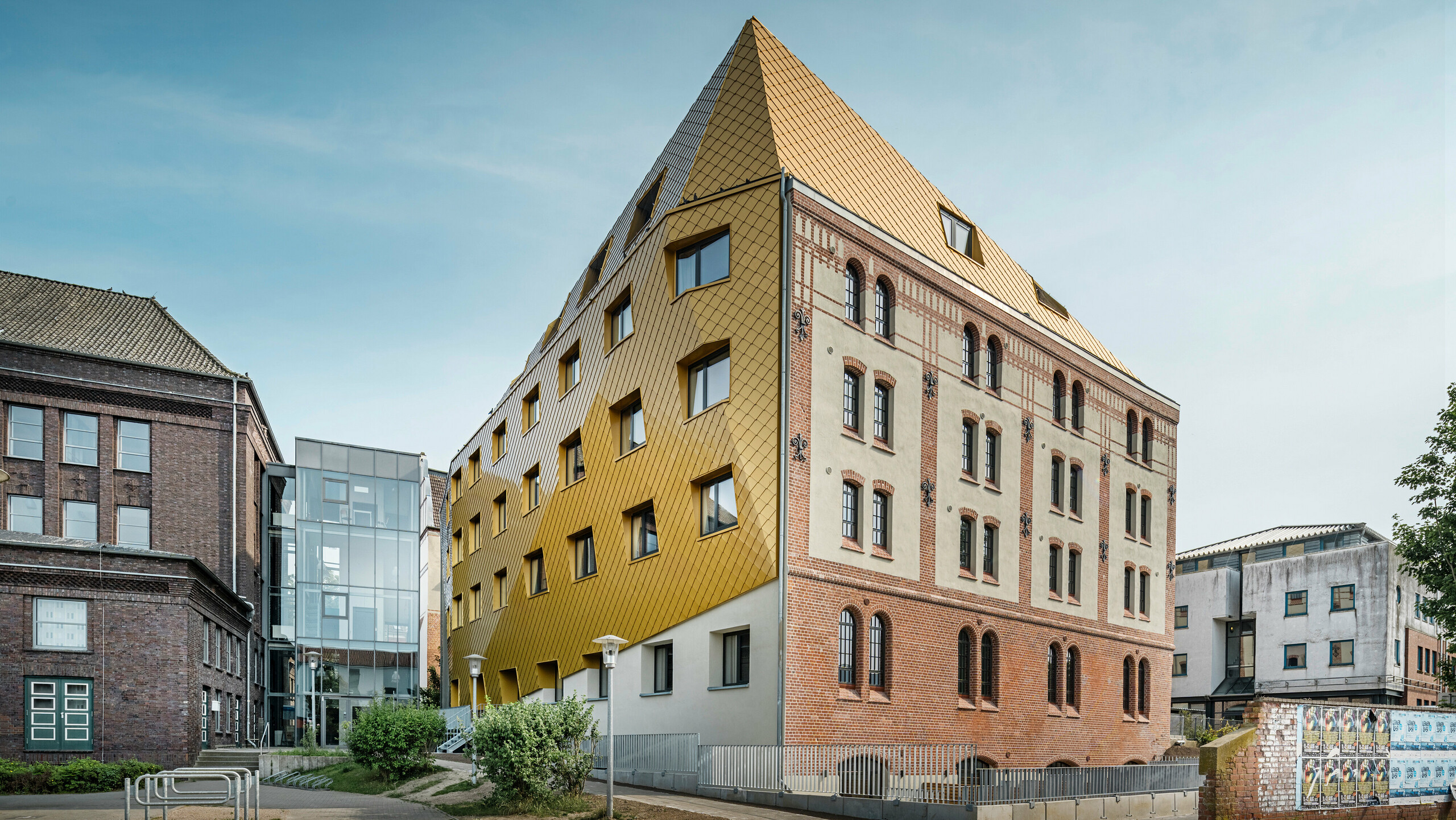 Bodenaufnahme auf "The Station" in Kiel. Das Studentenwohnheim ist schräg von der Seite zu sehen. Auf dem Bild wird der eklatante Kontrast zwischen der Aluminiumfassade aus PREFA Dach- und Wandrauten 29 × 29 in Mayagold auf der linken Seite und der denkmalgeschützten Putz- und Ziegelfassade auf der rechten Seite deutlich.