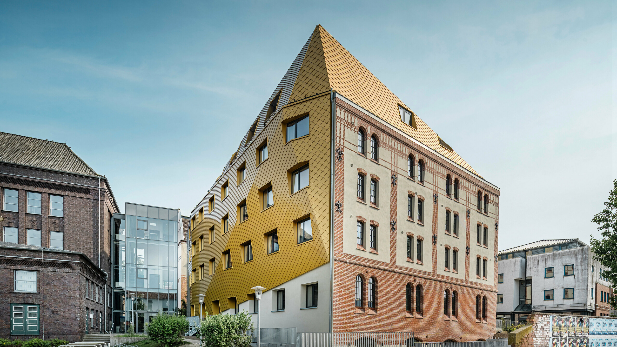 Bodenaufnahme auf "The Station" in Kiel. Das Studentenwohnheim ist schräg von der Seite zu sehen. Auf dem Bild wird der eklatante Kontrast zwischen der Aluminiumfassade aus PREFA Dach- und Wandrauten 29 × 29 in Mayagold auf der linken Seite und der denkmalgeschützten Putz- und Ziegelfassade auf der rechten Seite deutlich.