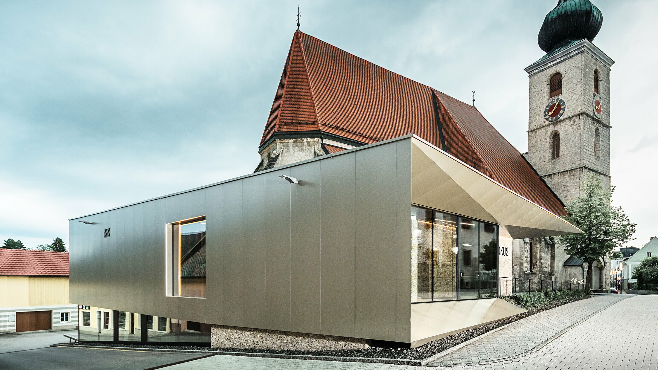 Die markante bronzefarbene Verbundplatten-Fassade des Pfarrzentrums in Sierning reckt sich um die kleine Kirche im Hintergrund