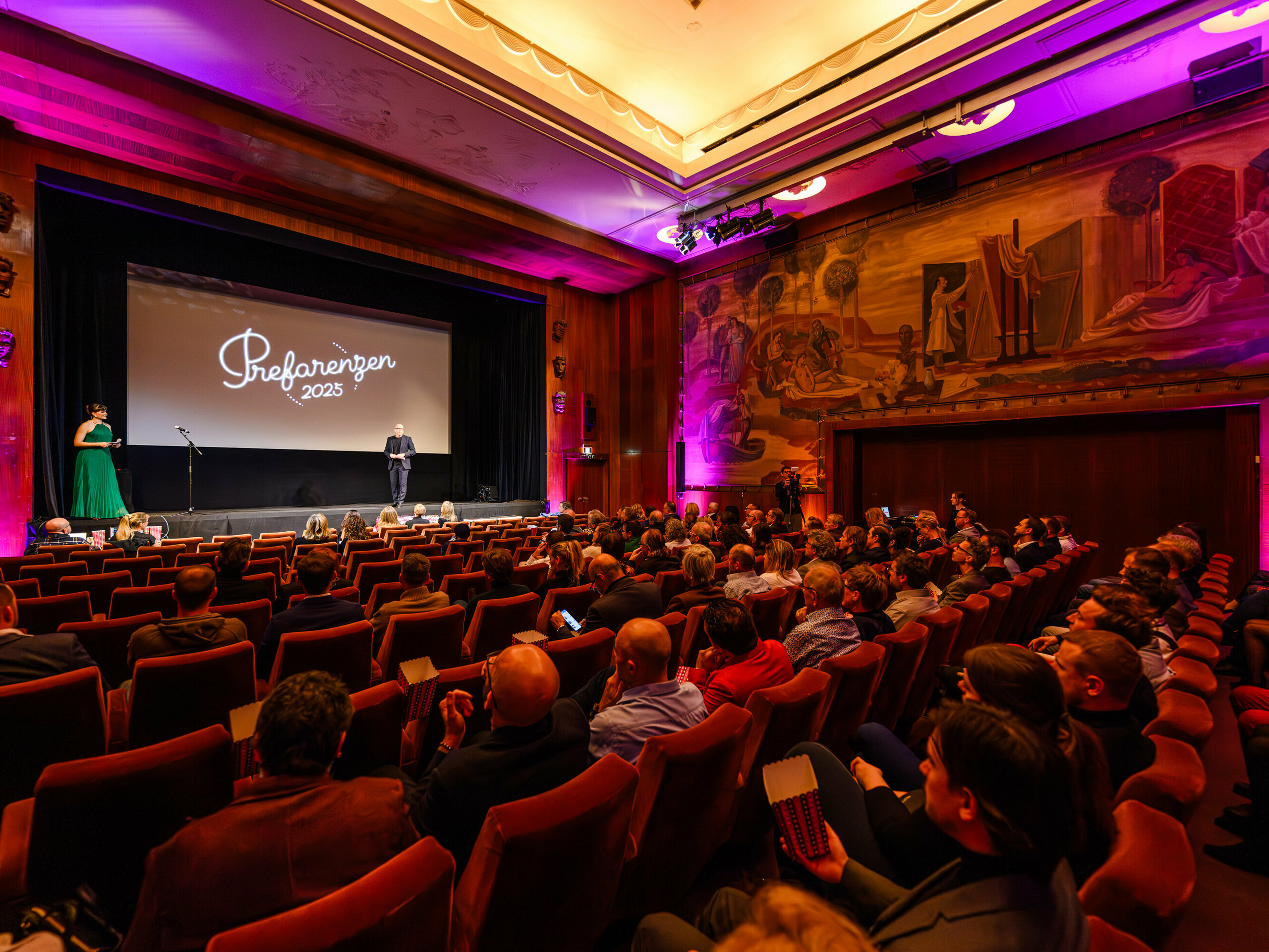 Das historische Stadtkino Wien während der PREFARENZEN-Premiere 2025: Jürgen Jungmair, Marketingleiter von PREFA, hält eine Rede vor einem internationalen Publikum aus Architektur-, Planungs- und Medienexpert:innen. Im Hintergrund ist das Logo ‚Prefarenzen 2025‘ auf der Leinwand zu sehen, umrahmt von stilvoller Beleuchtung und beeindruckender Wandkunst.