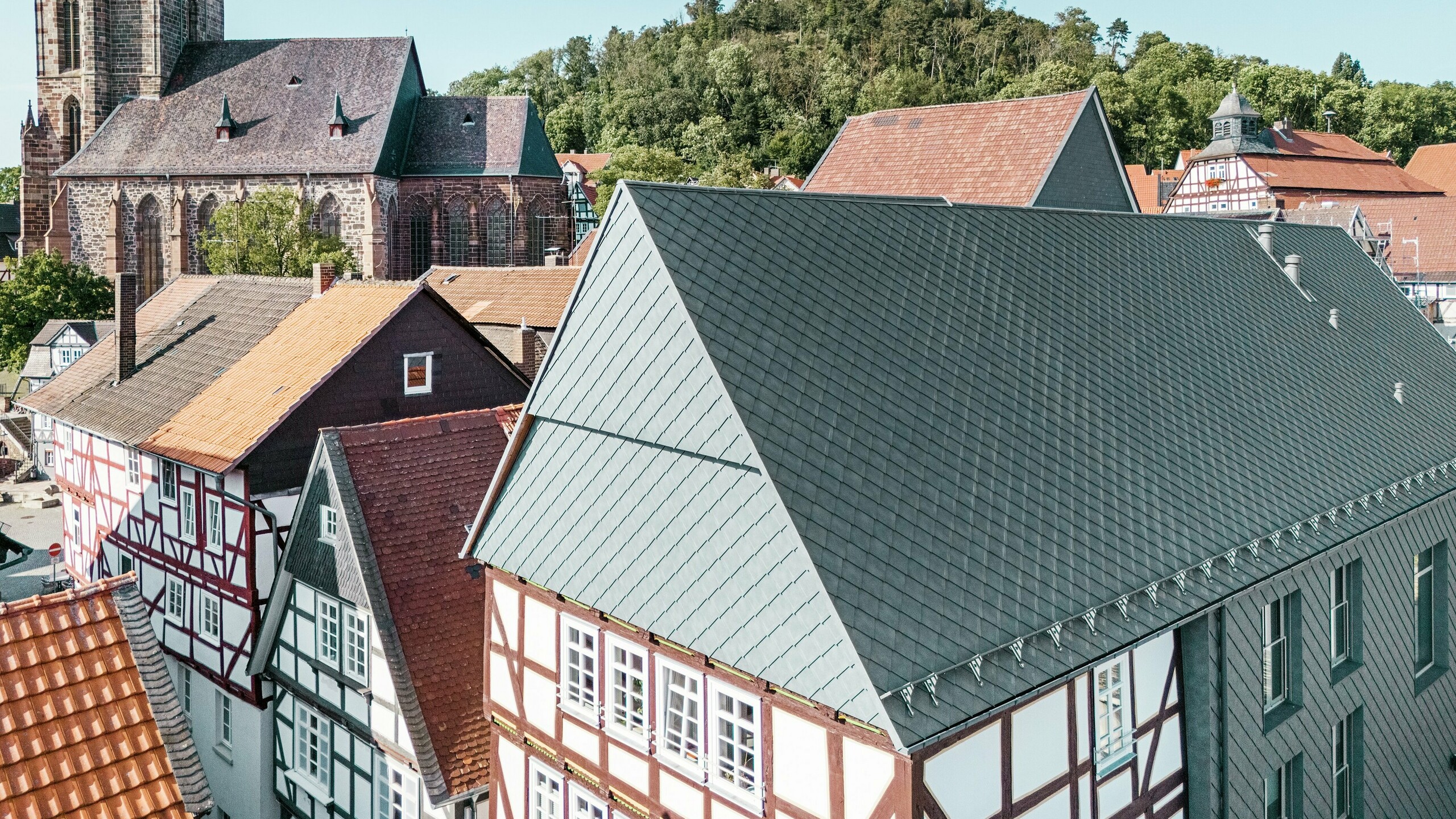 Das Dach des sanierten Volkshauses in Homberg, DE, mit PREFA Aluminiumrauten 29 × 29 in P.10 Steingrau, eingebettet in die historische Altstadt mit Fachwerkhäusern, Kirche und Burg. Die langlebige und robuste Aluminiumverkleidung verbindet modernes Design mit dem Charme der historischen Umgebung. In der Bildmitte ist der Giebel des Gebäudes zu sehen, im Hintergrund die Kirche und der Burgberg. 