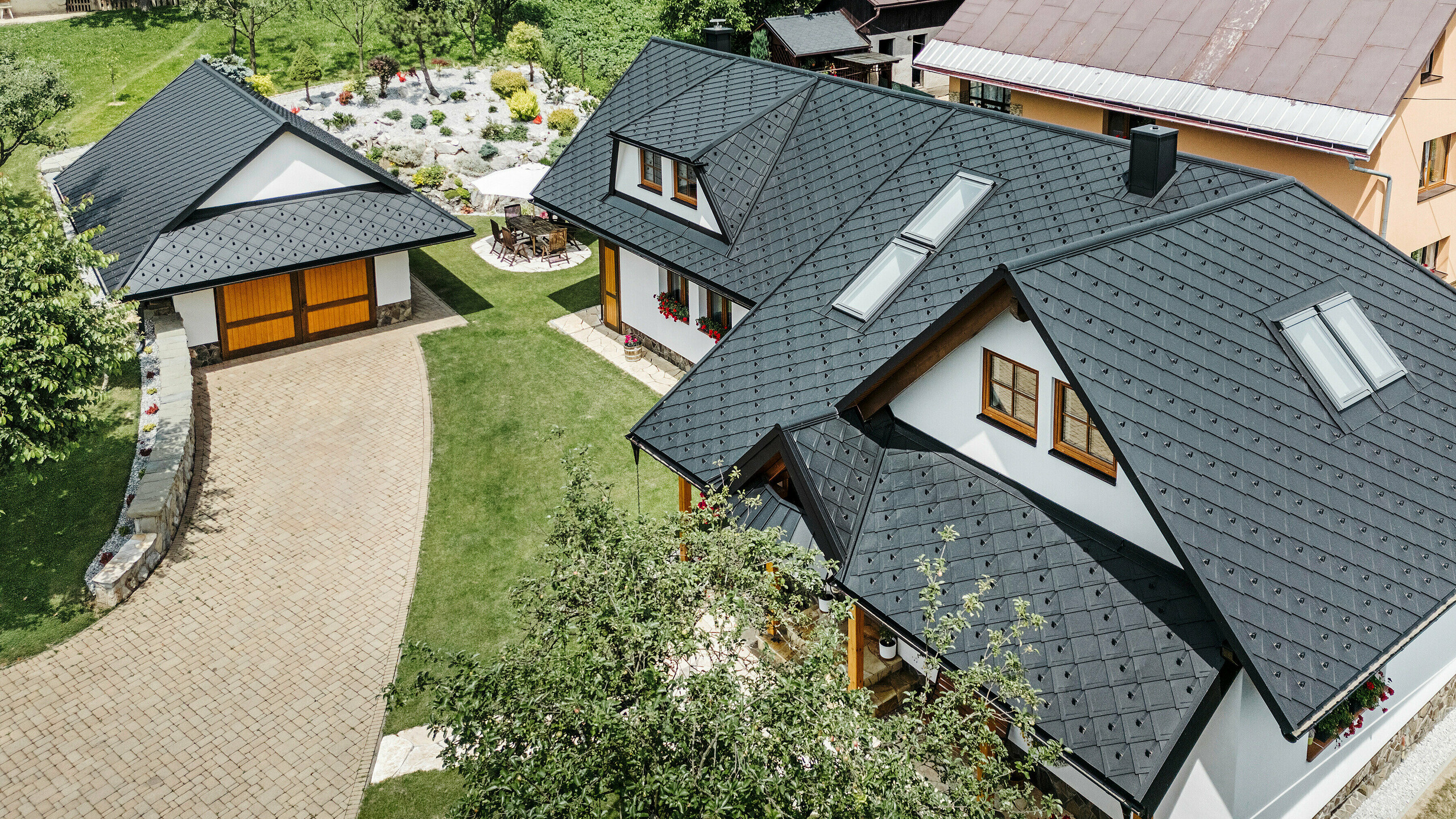 Vogelperspektive auf ein Einfamilienhaus in Stara Bystrica, Slowakei, mit einem Dach aus PREFA Dachrauten 29 × 29 in P.10 Schwarz. Das Dach, ausgeführt in gleichmäßiger Rautenoptik, umfasst Dachschrägenfenster und Trapezgauben auf beiden Seiten. Die Holzverzierungen an den Fenstern sowie die farbenfrohen Blumenarrangements verleihen dem Haus ein einladendes und harmonisches Erscheinungsbild. Der gepflegte Vorgarten und die gepflasterte Einfahrt ergänzen das Gesamtbild perfekt.